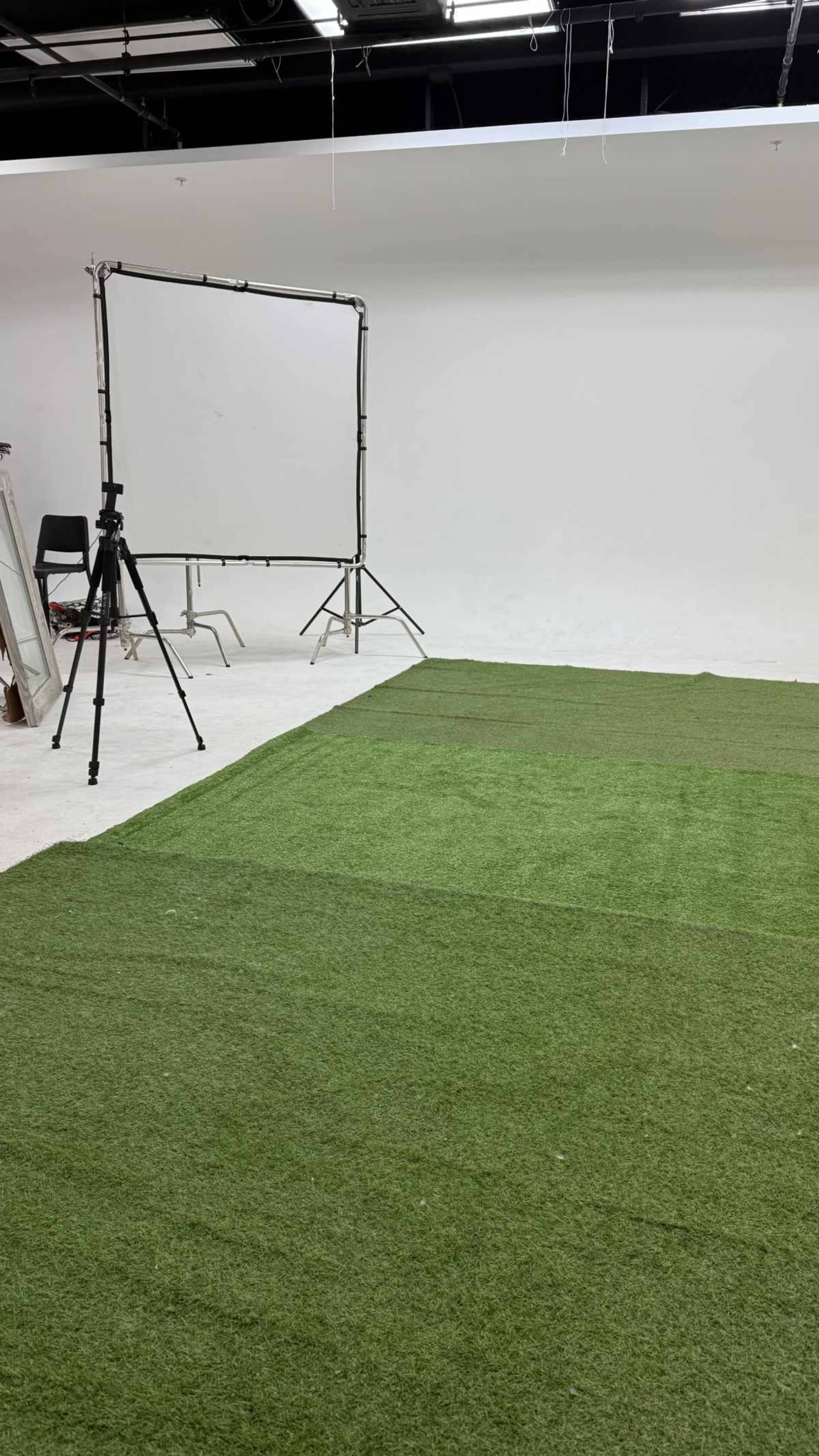 The image shows a studio space with artificial grass flooring, a backdrop stand, and a tripod set up against a white wall.