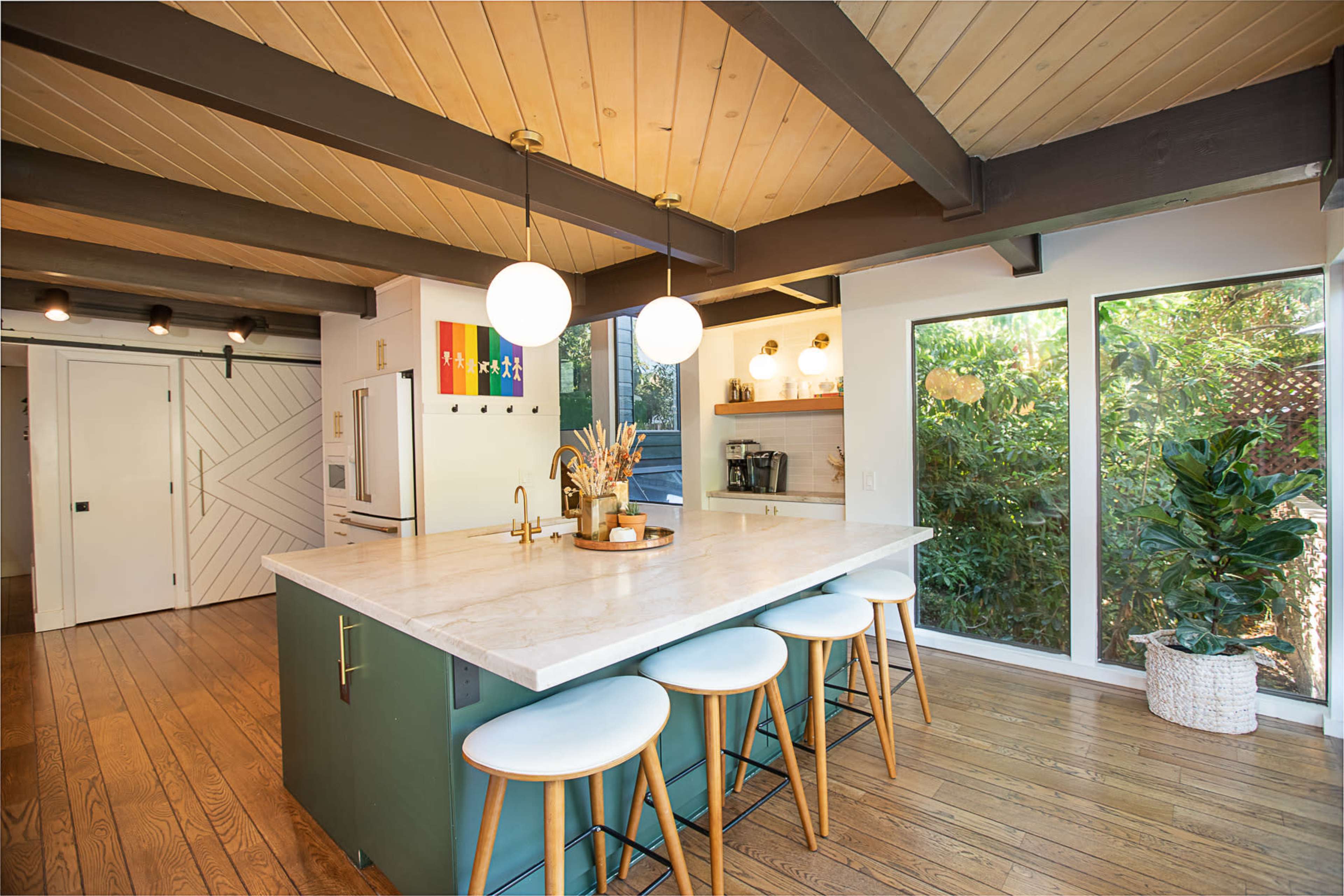 A modern kitchen features a marble island with seating, large windows displaying greenery, and wooden beams on the ceiling.
