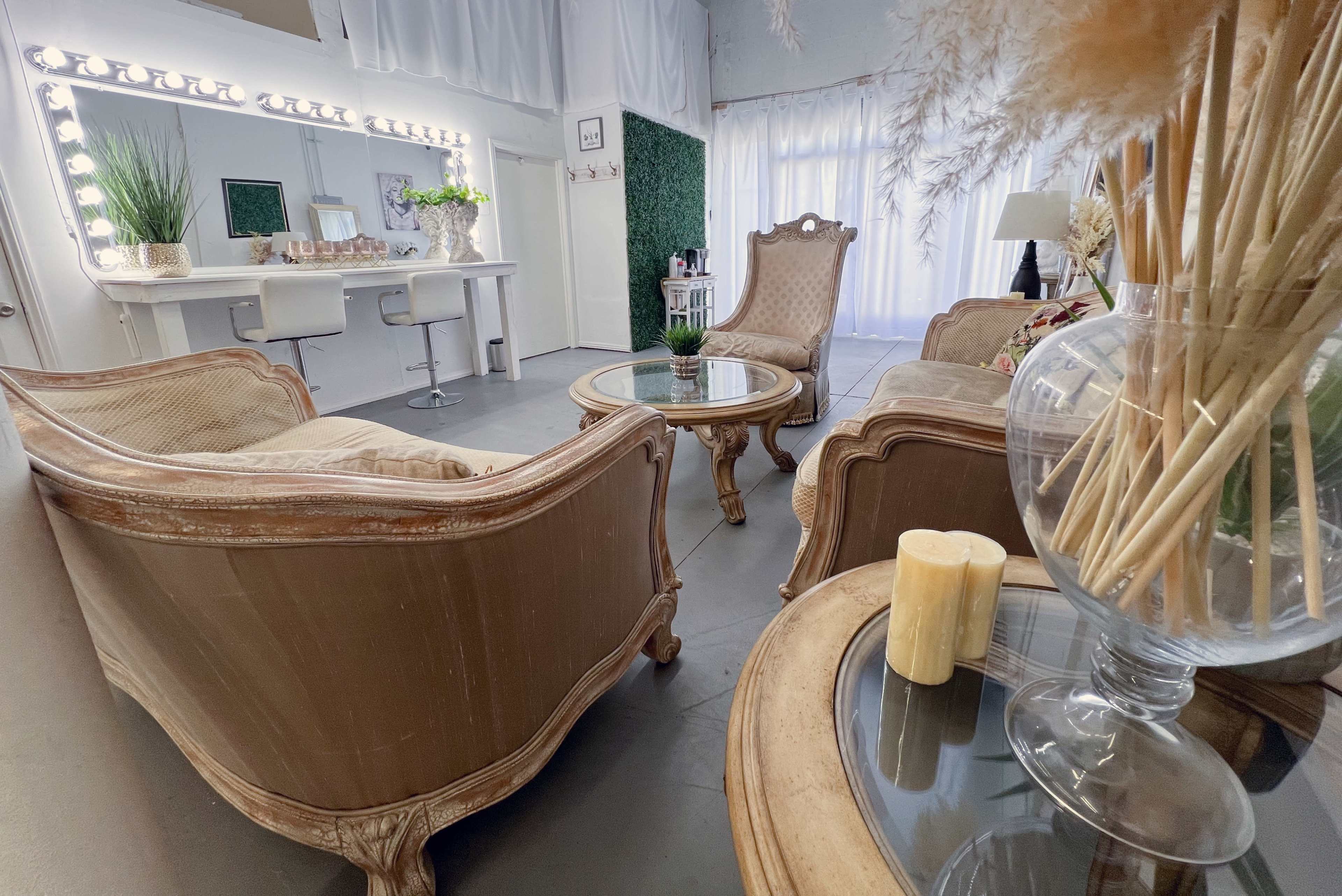 The image shows a stylish interior featuring two antique-style chairs, a glass table, and a makeup vanity with bright lights, all set in a spacious room with light curtains and decorative plants.