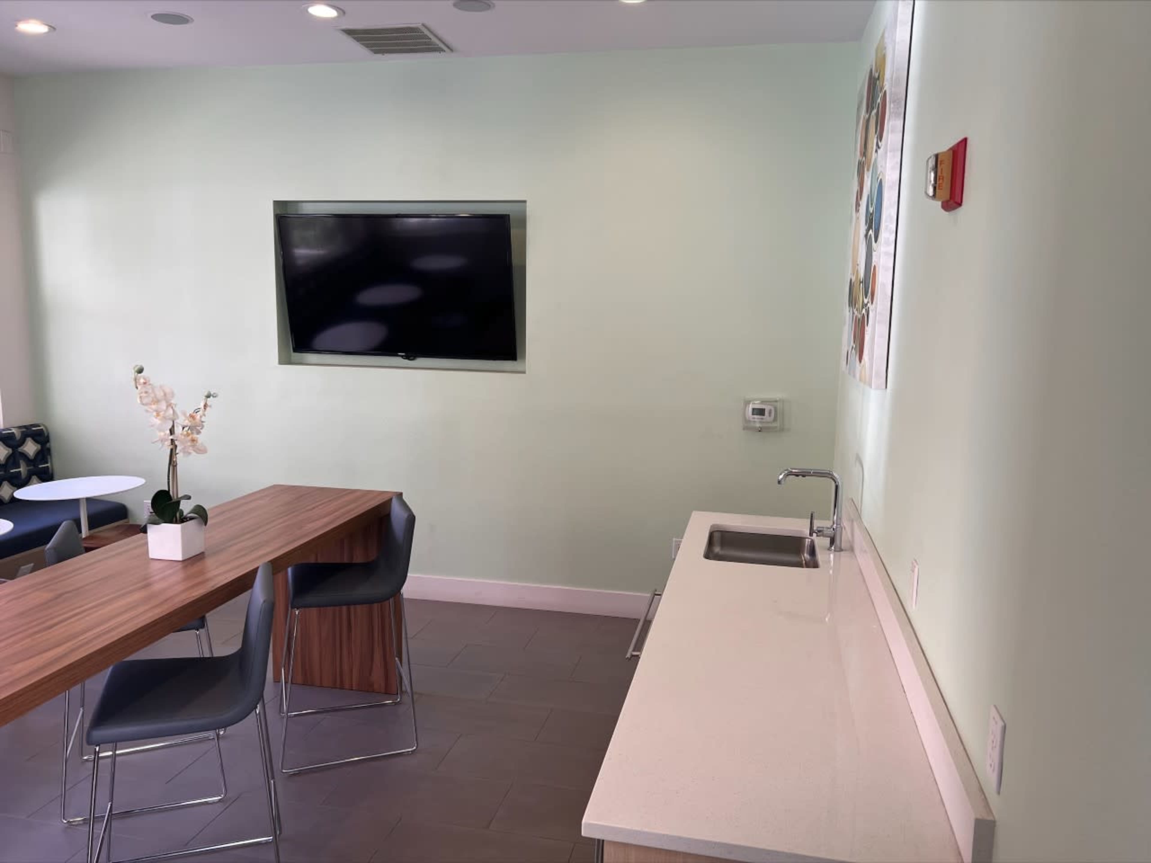 A modern kitchen area with a sink, a table, and chairs, along with a wall-mounted TV and light green walls.