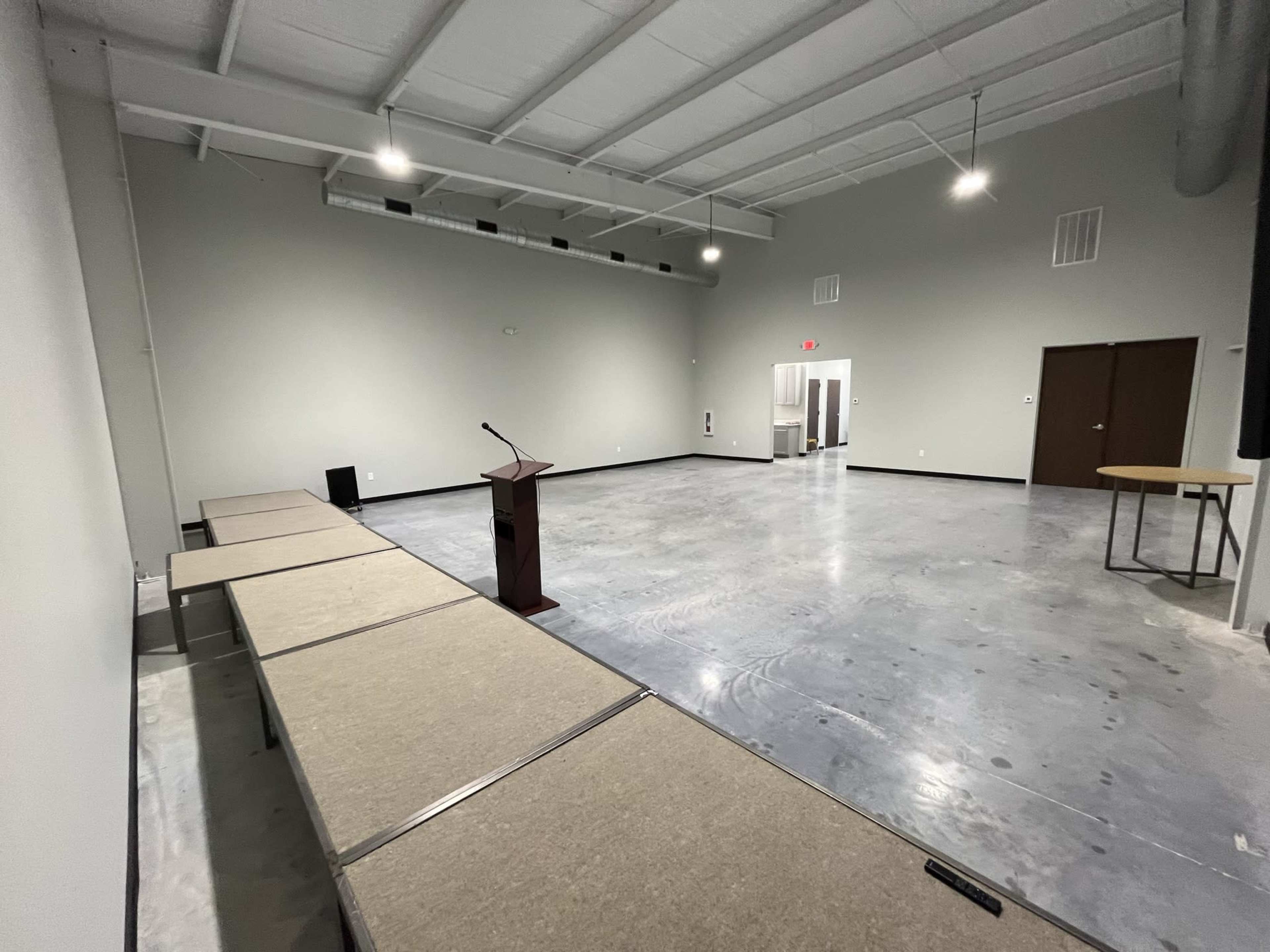 A large, empty room with gray walls, concrete floors, a podium at the front, and several folding tables arranged in a U-shape.
