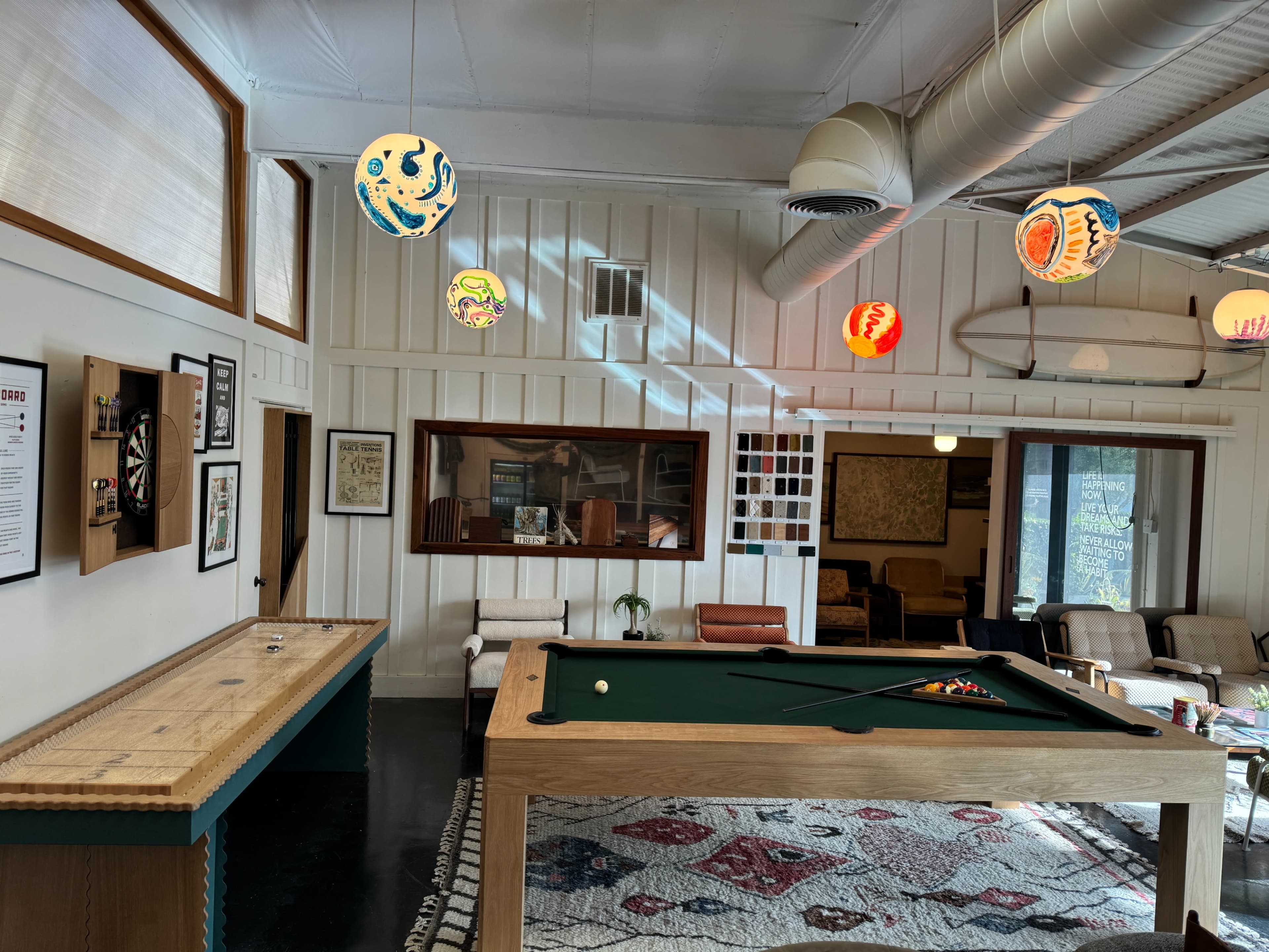 A recreational room with a green billiards table, decorative overhead lights, and a seating area featuring armchairs and a large window.