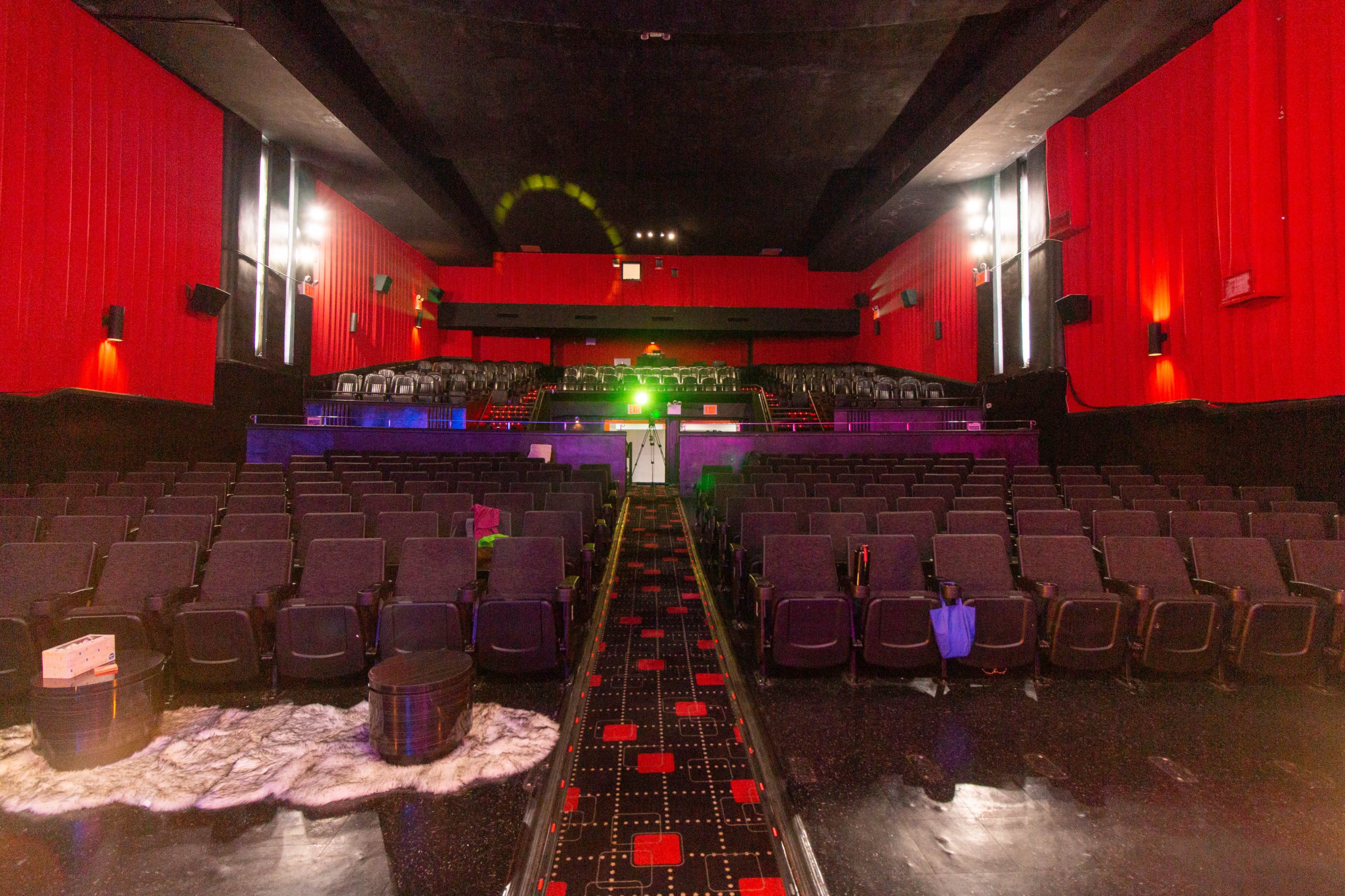 The interior of an empty theater with red walls, rows of black seats, and a central aisle leading to a stage area illuminated by a green light.