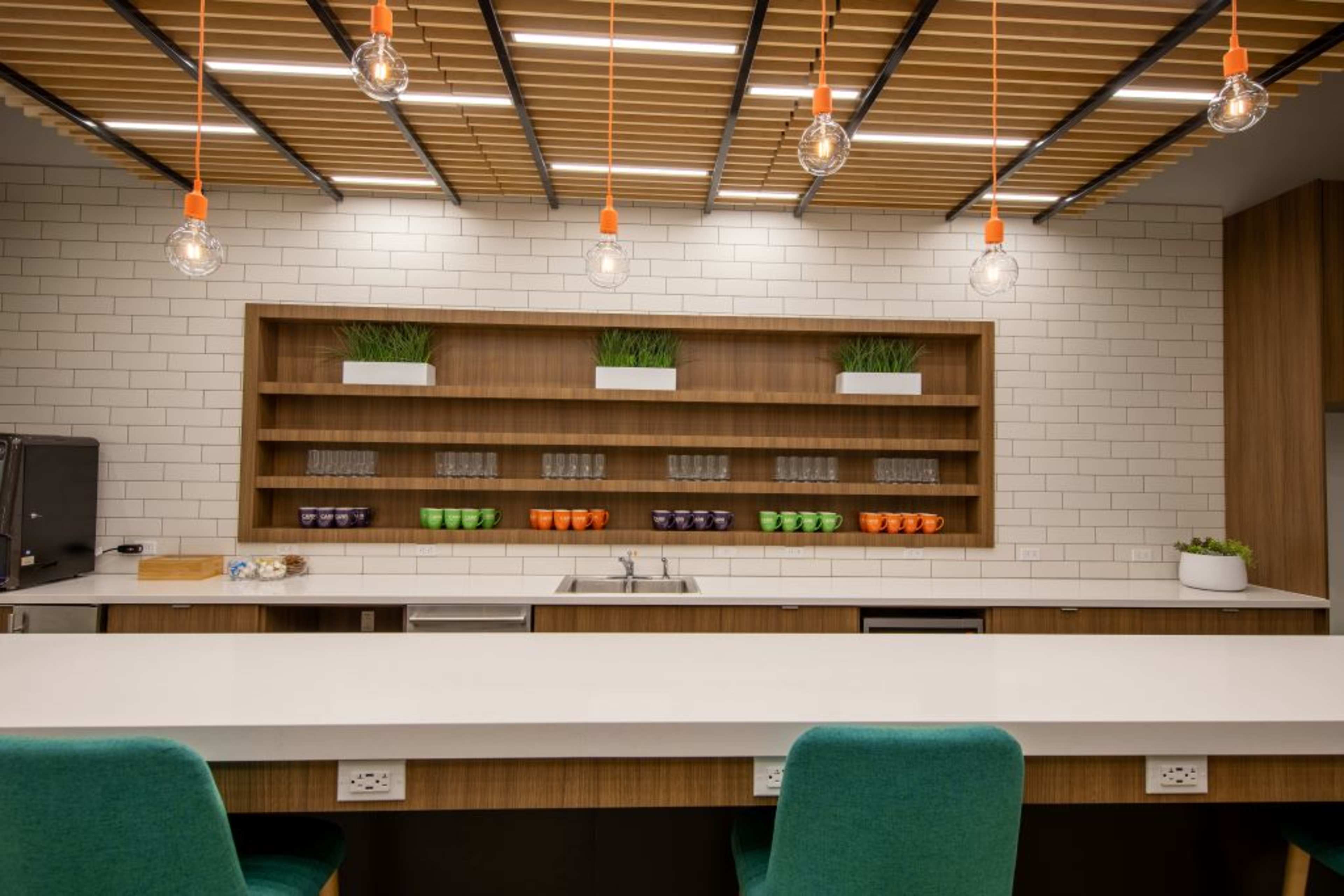 A modern kitchen features a long countertop, overhead lighting, wooden shelves with plants and colored glassware, and a sink against a tiled wall.