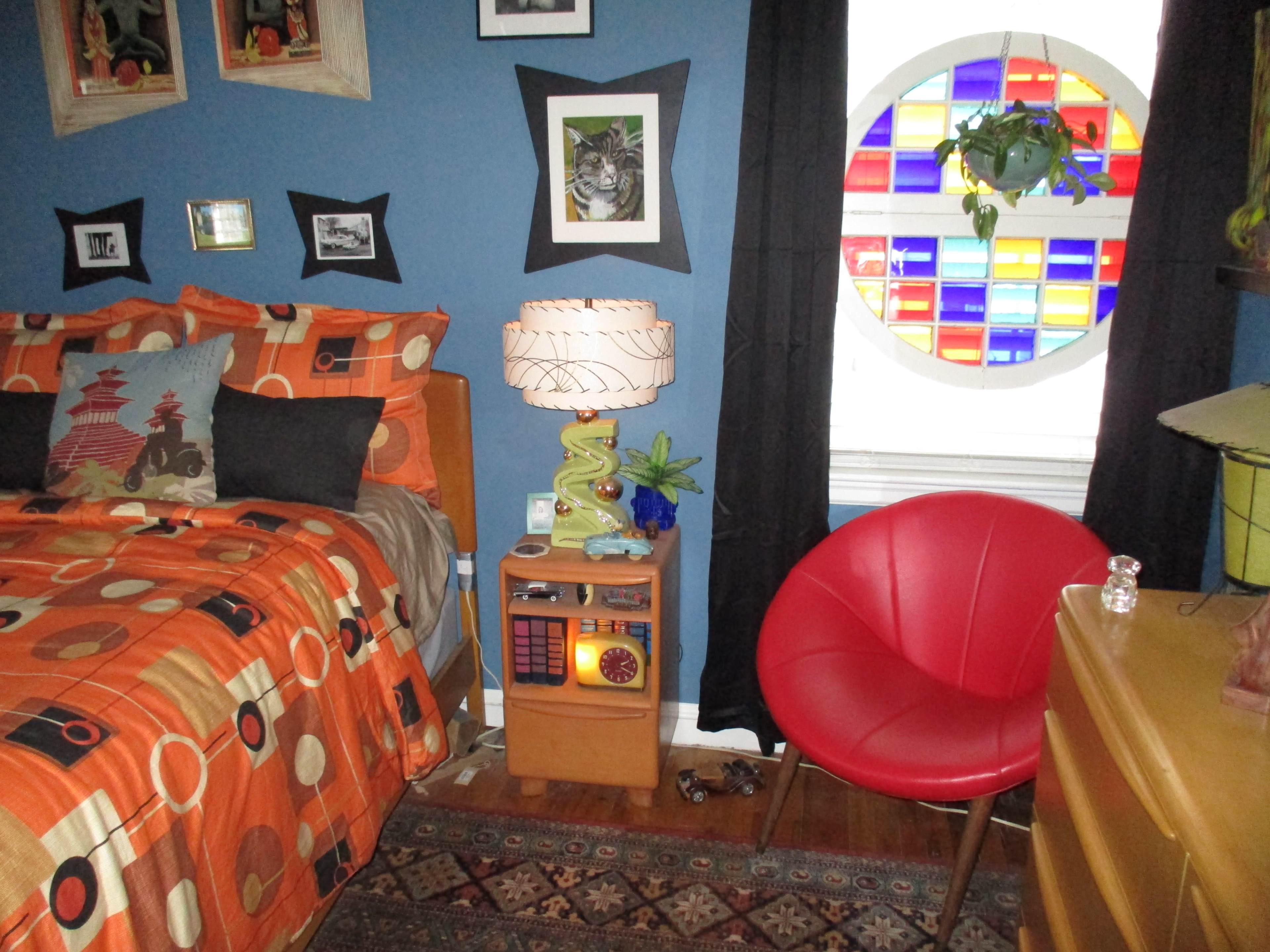 A colorful bedroom featuring a bed with patterned bedding, a round stained glass window, a red chair, and various decorative items.
