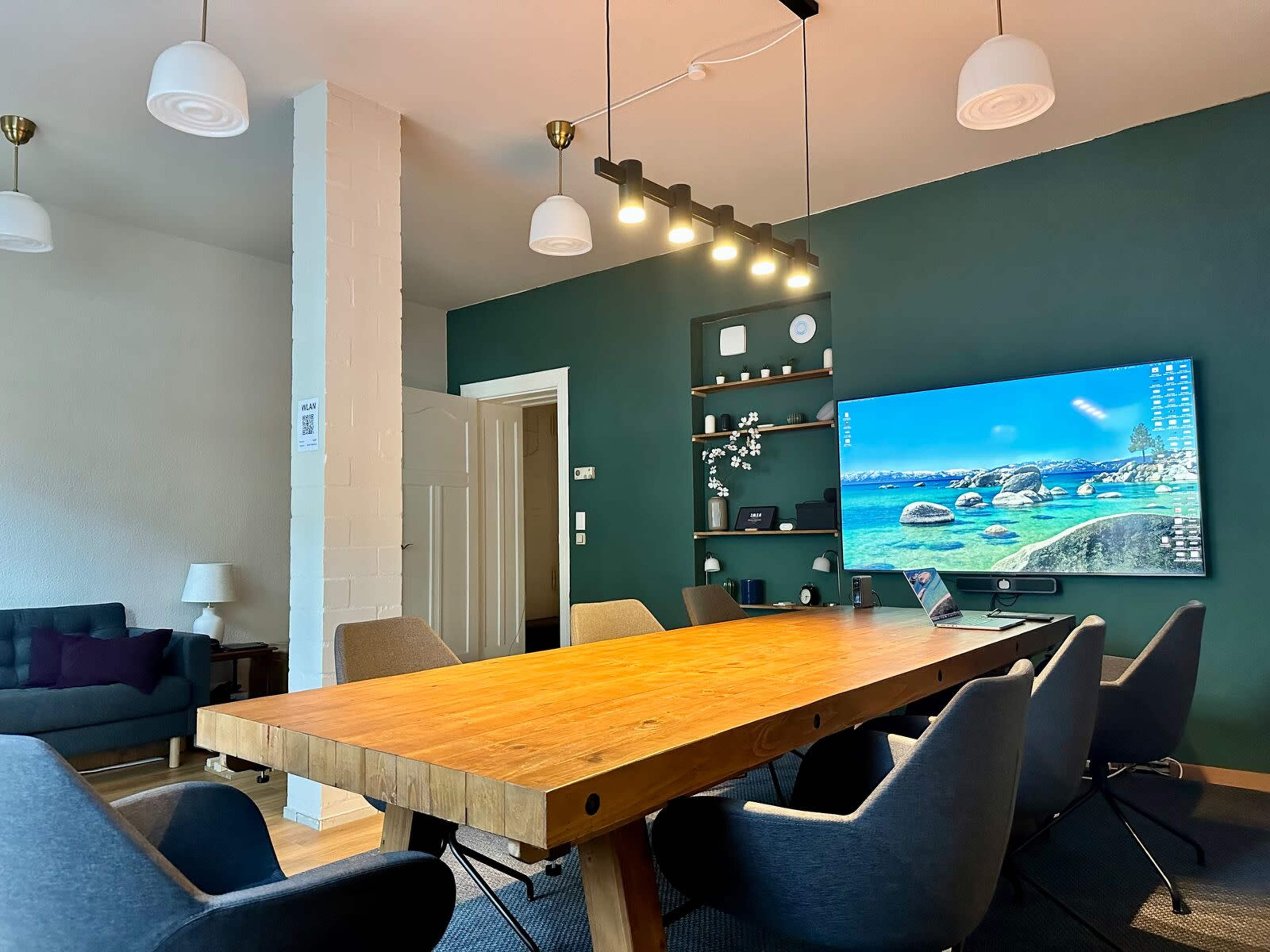 The image shows a modern conference room with a large wooden table surrounded by chairs, a mounted television displaying a scenic background, and a dark green accent wall.