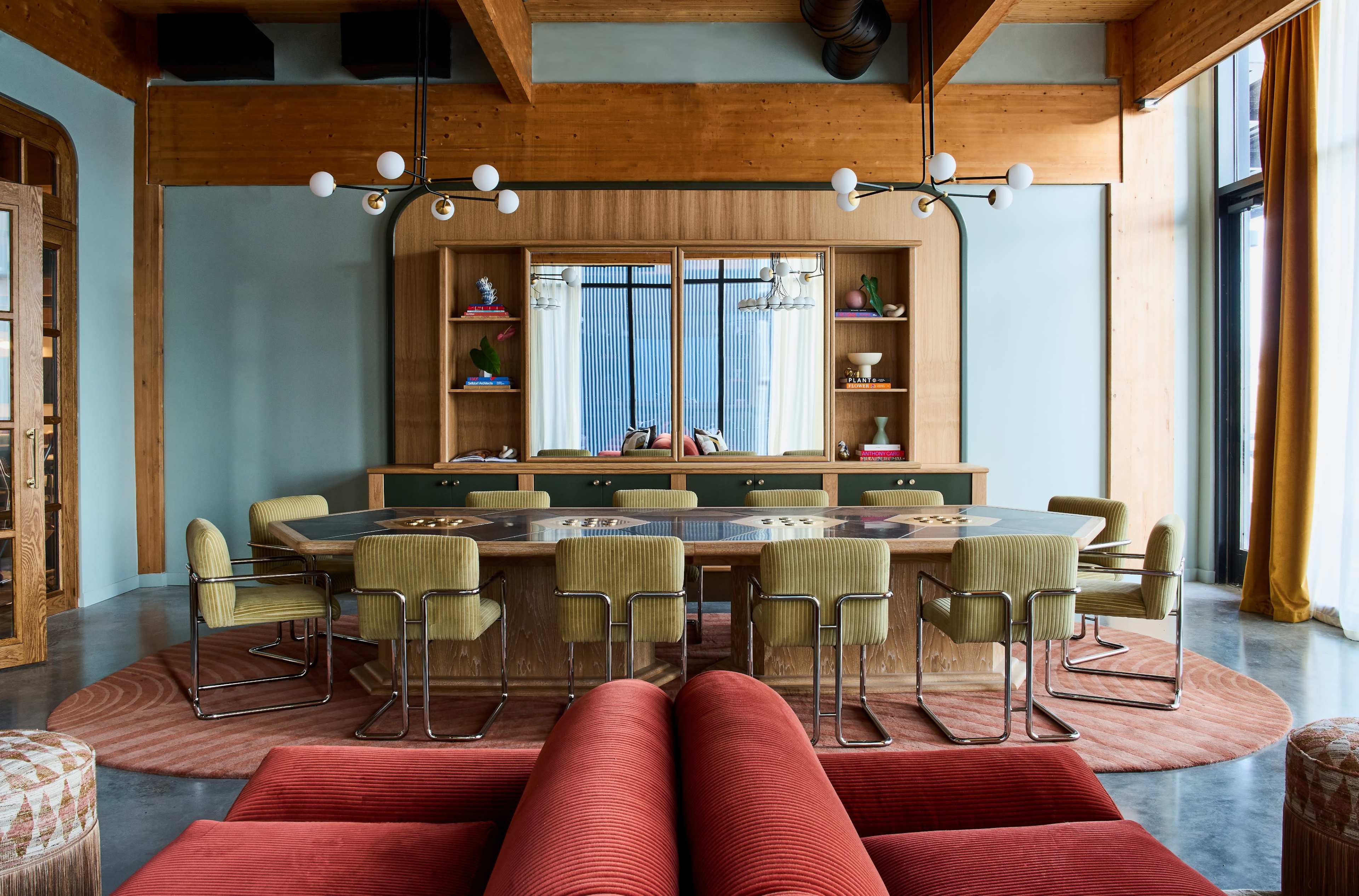 A modern, spacious dining area featuring a large central table surrounded by upholstered chairs, complemented by a wooden wall unit and large windows.
