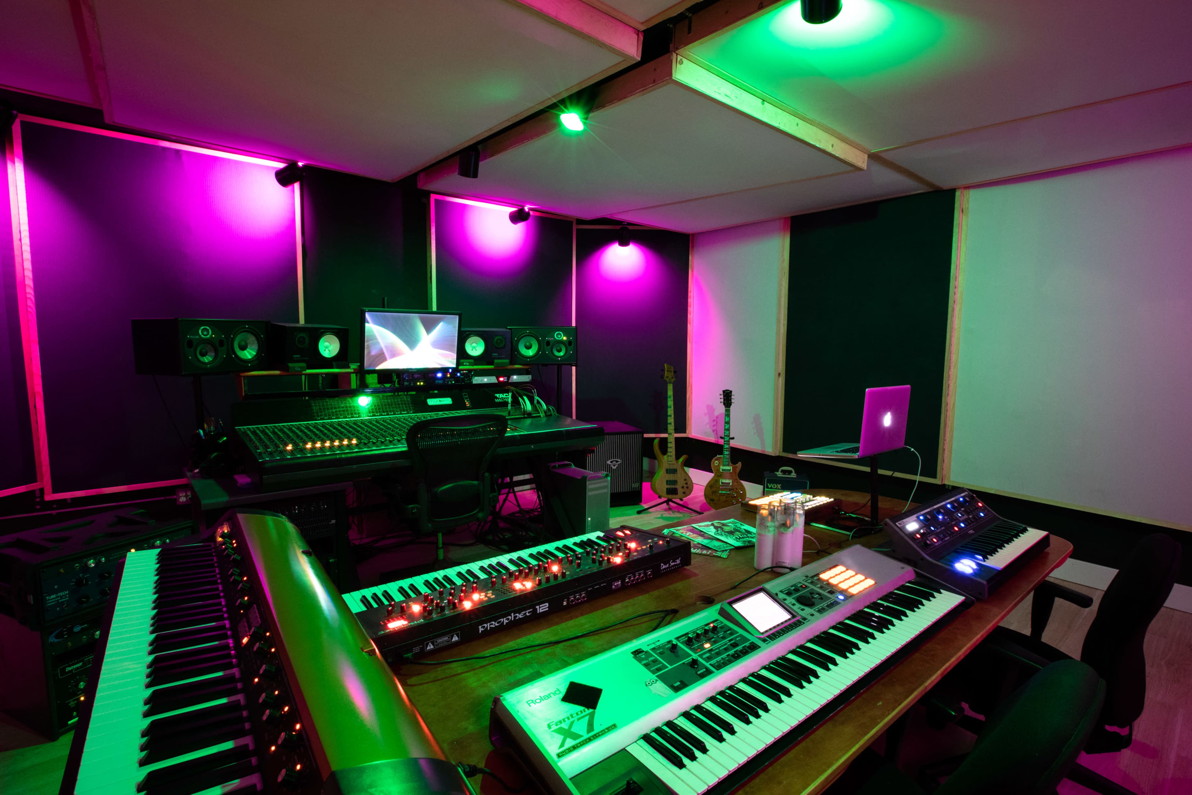 A music production studio featuring multiple electronic instruments, sound monitors, a computer setup, and colorful ambient lighting.