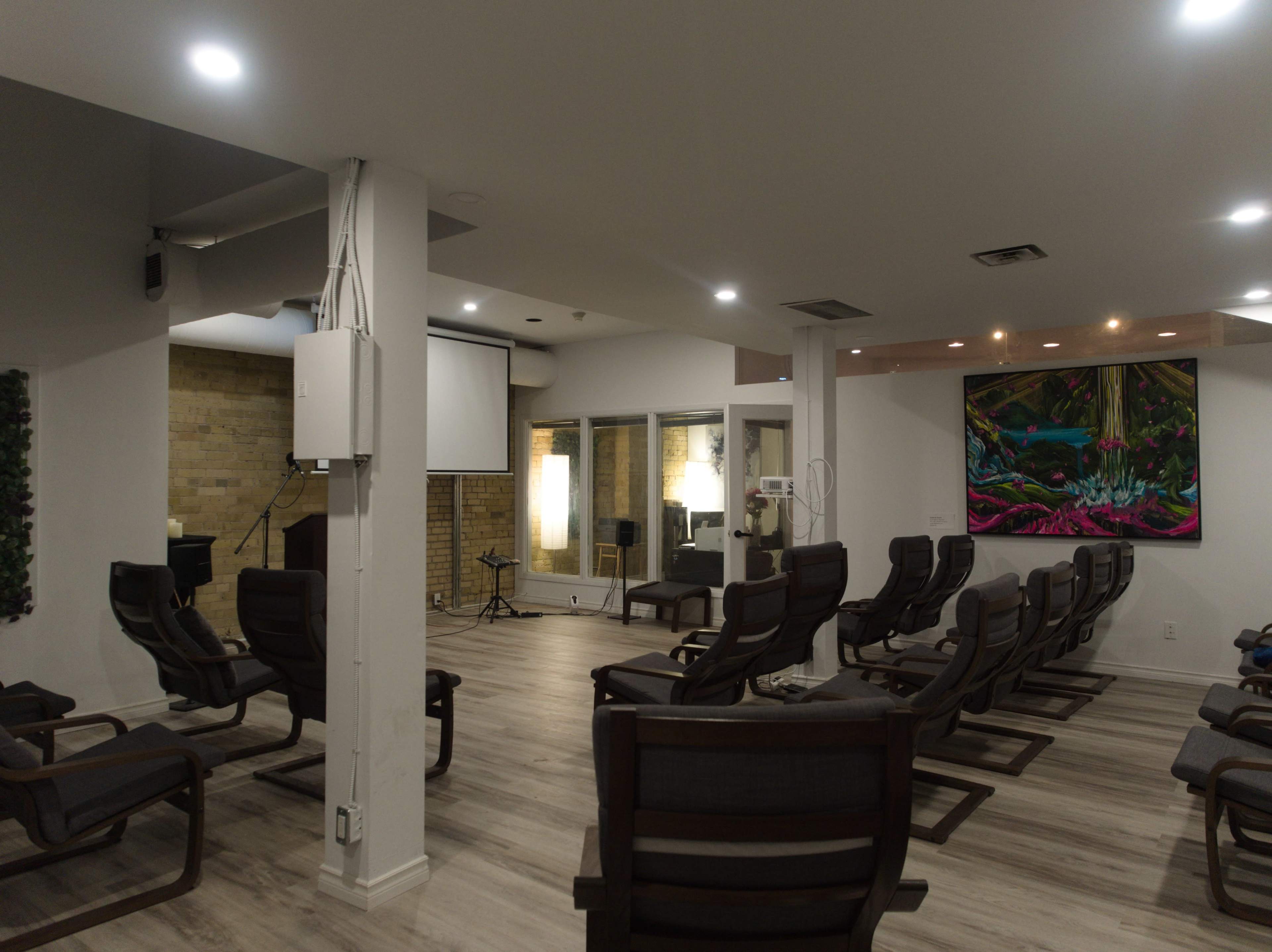 The image shows a spacious room with several rows of chairs, a large screen, and a vibrant painting on the wall.