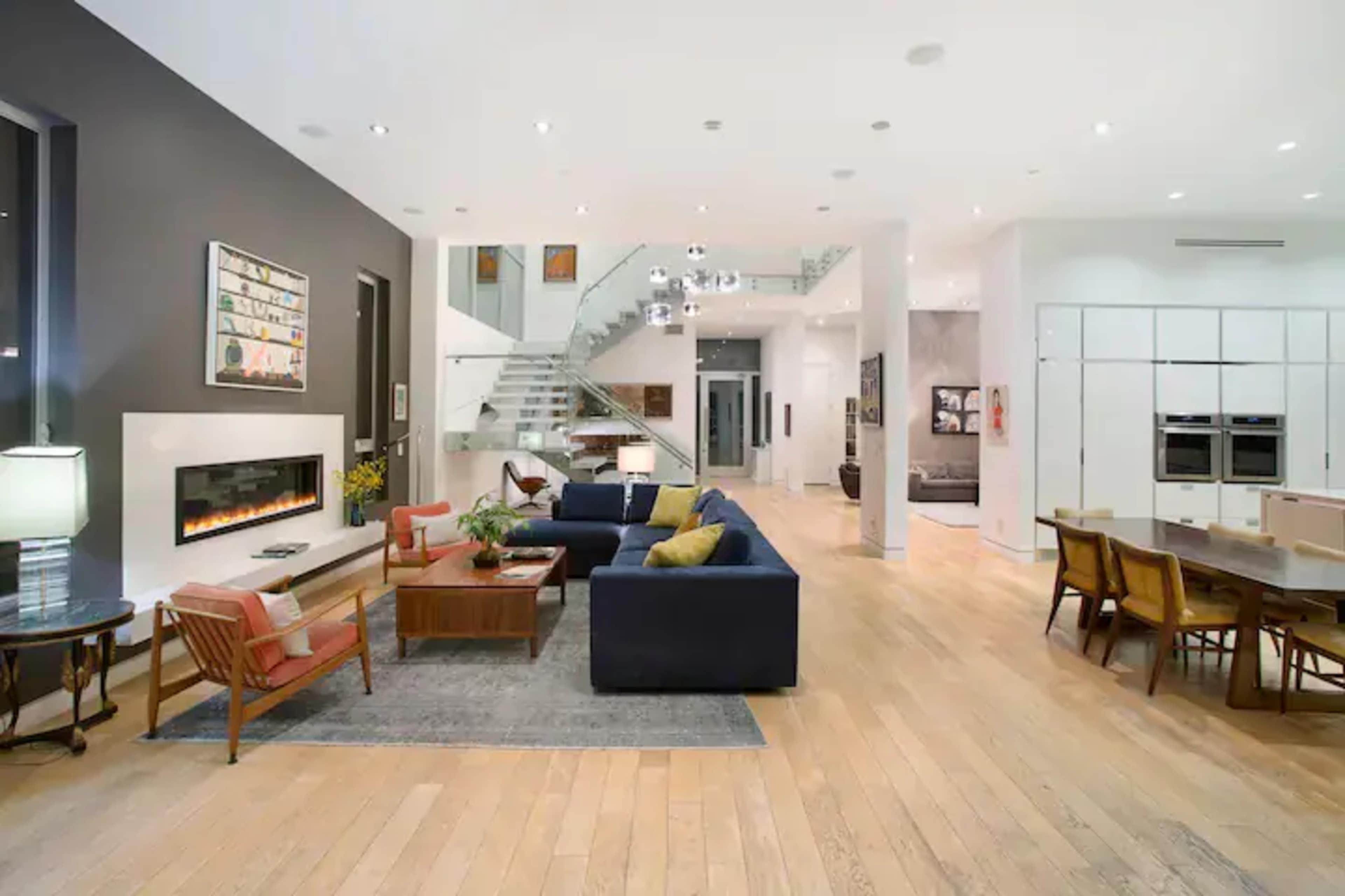 A spacious modern living room and dining area with hardwood floors, a fireplace, and a staircase leading to an upper level.