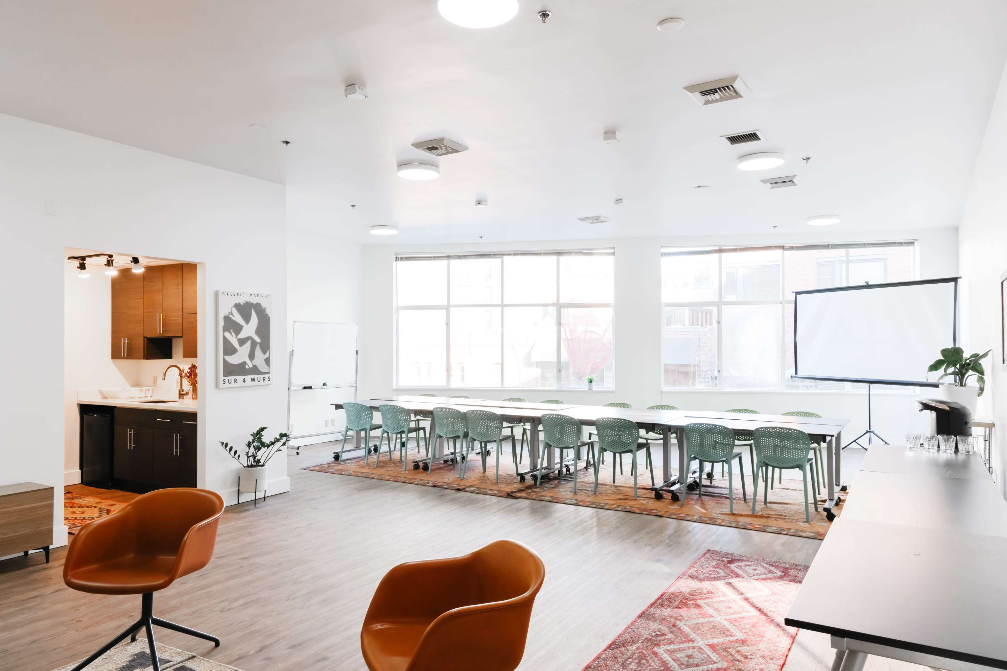 A modern conference room with a long table surrounded by chairs, a large screen, and a kitchenette visible in the background.