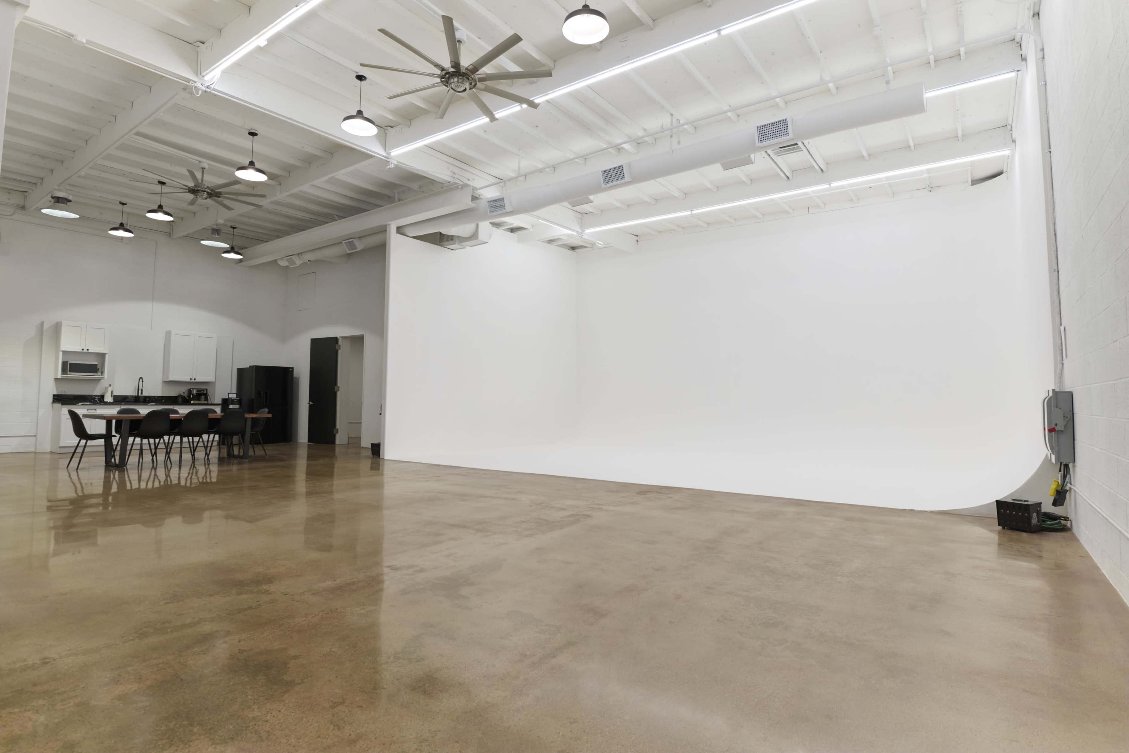 The image shows a spacious, well-lit studio with a clean white backdrop, polished concrete floor, and a kitchen area with a dining table and chairs in the corner.