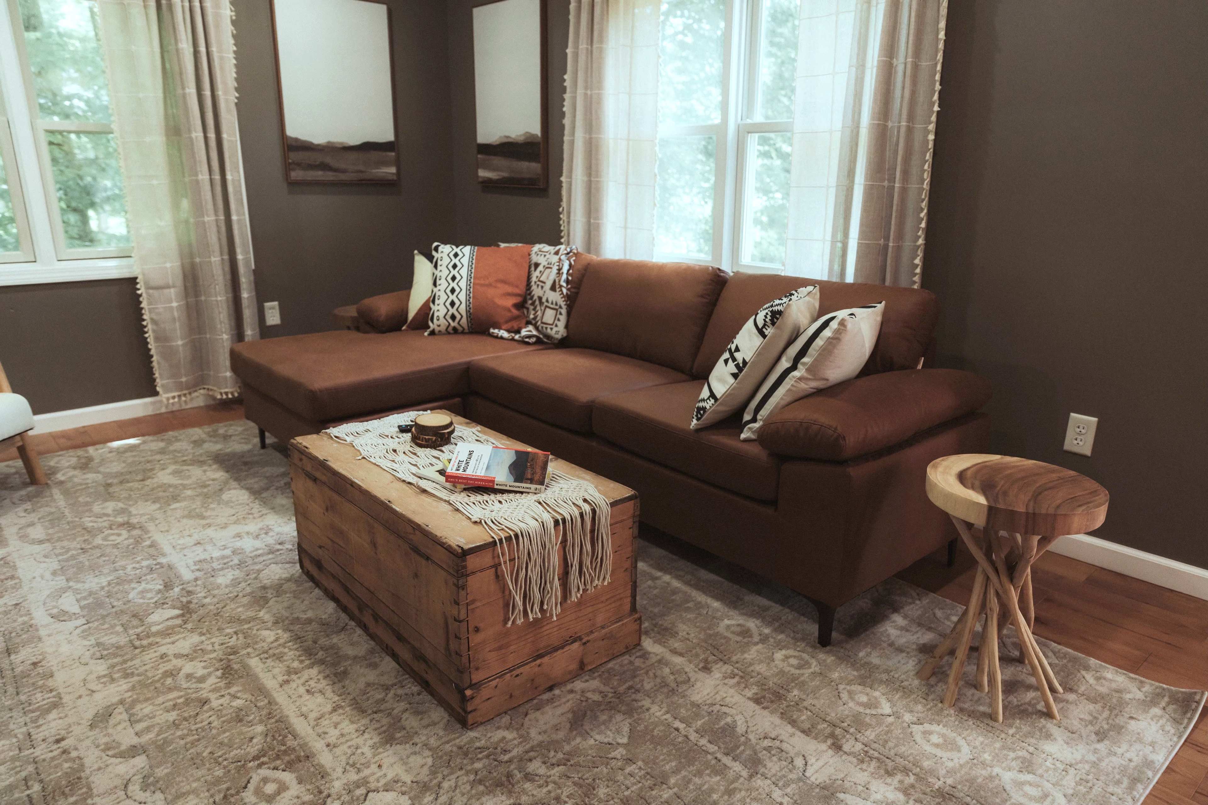 A brown sectional sofa with patterned pillows and a wooden coffee table in a cozy living room with large windows dressed in sheer curtains.