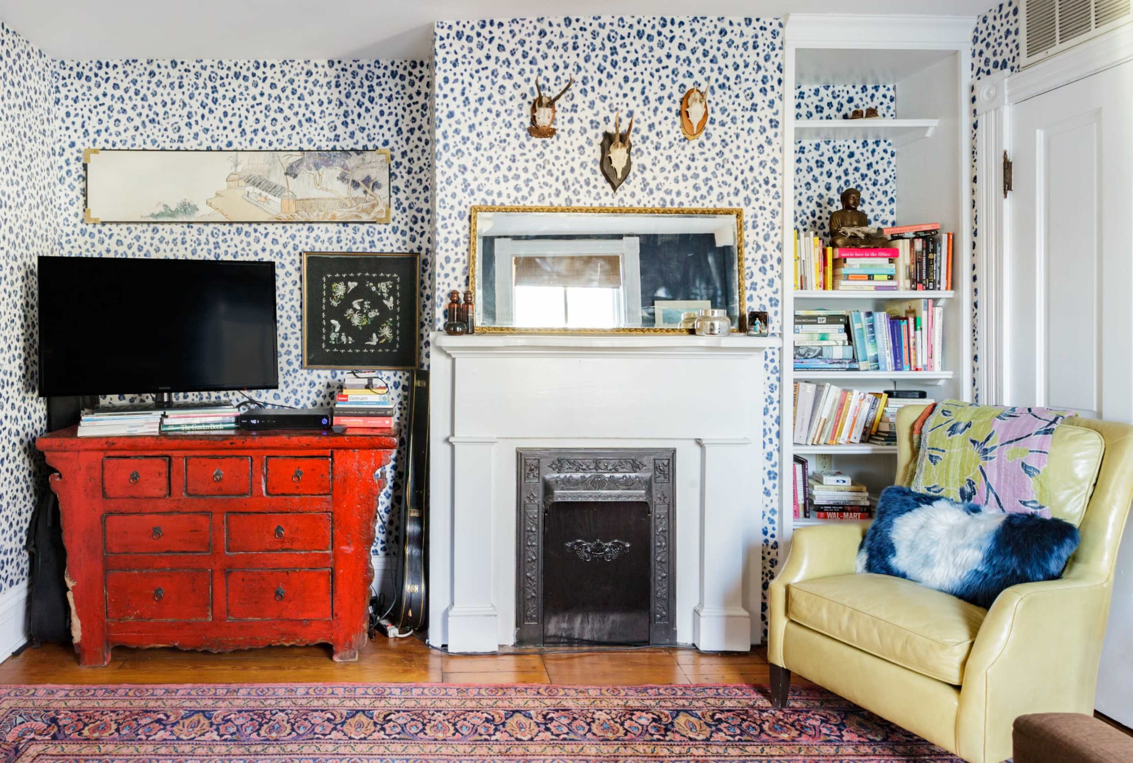 A cozy living room featuring a red dresser, a television, a white fireplace, a bookshelf filled with books, and a yellow armchair against a patterned wallpaper.