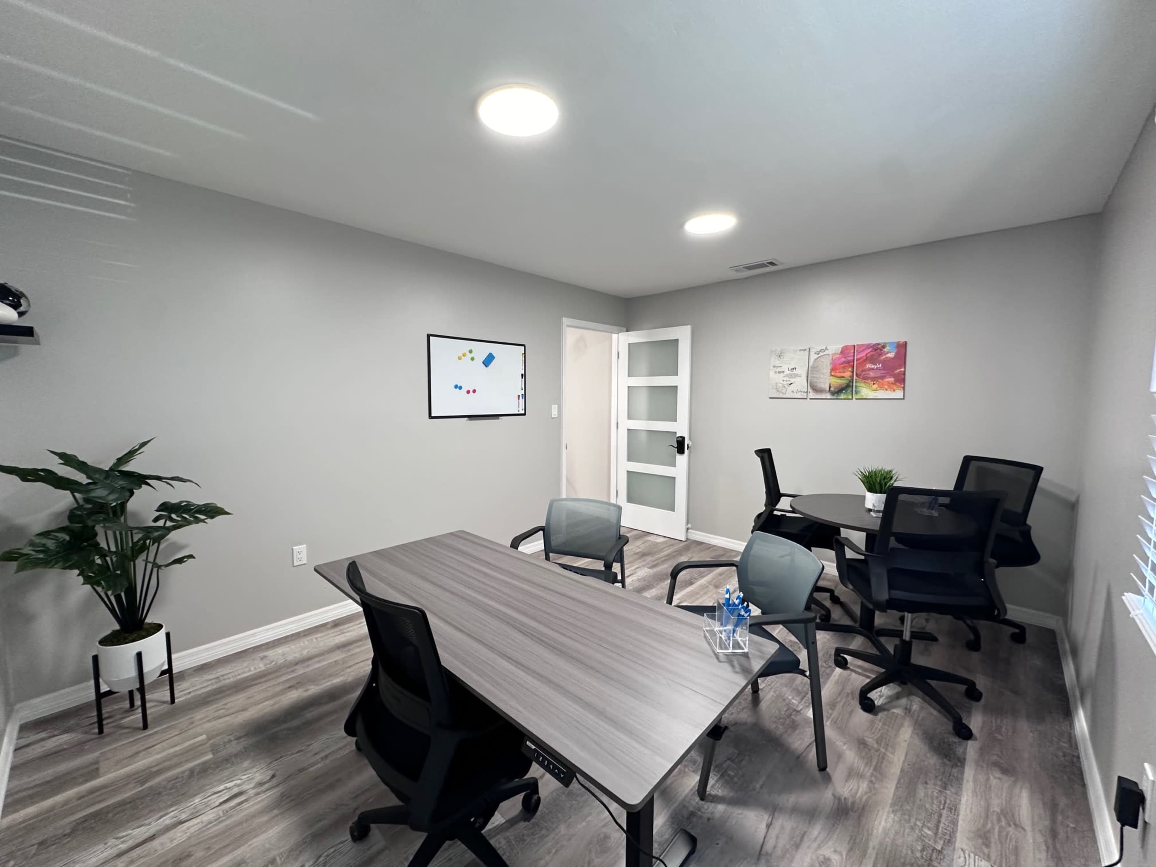 The image shows a modern office space with a large table, several chairs, a plant, and a whiteboard on the wall.
