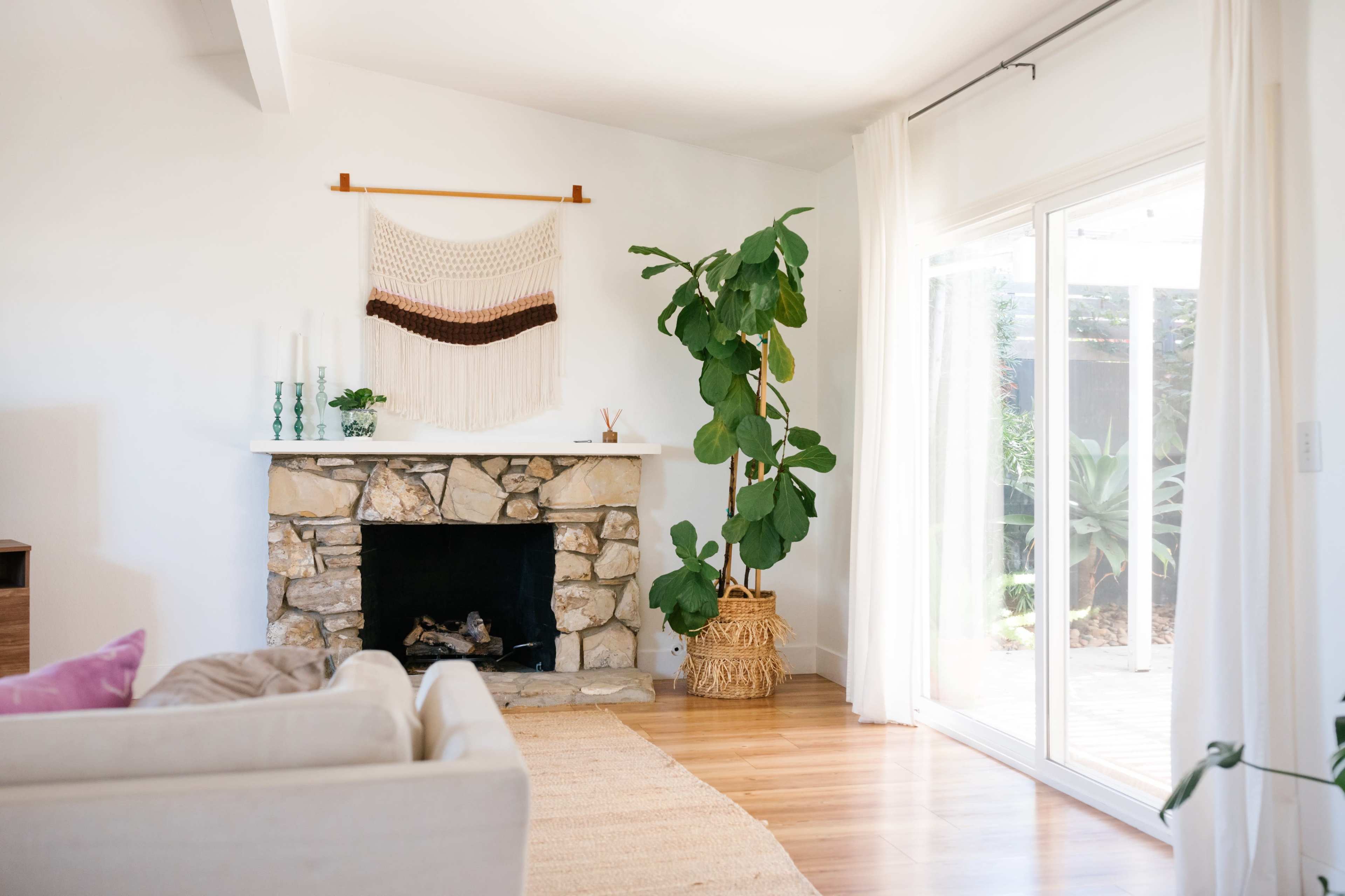 A living room features a stone fireplace with a macramé wall hanging above it, a large plant in a woven basket, and sliding glass doors that lead to an outdoor area.