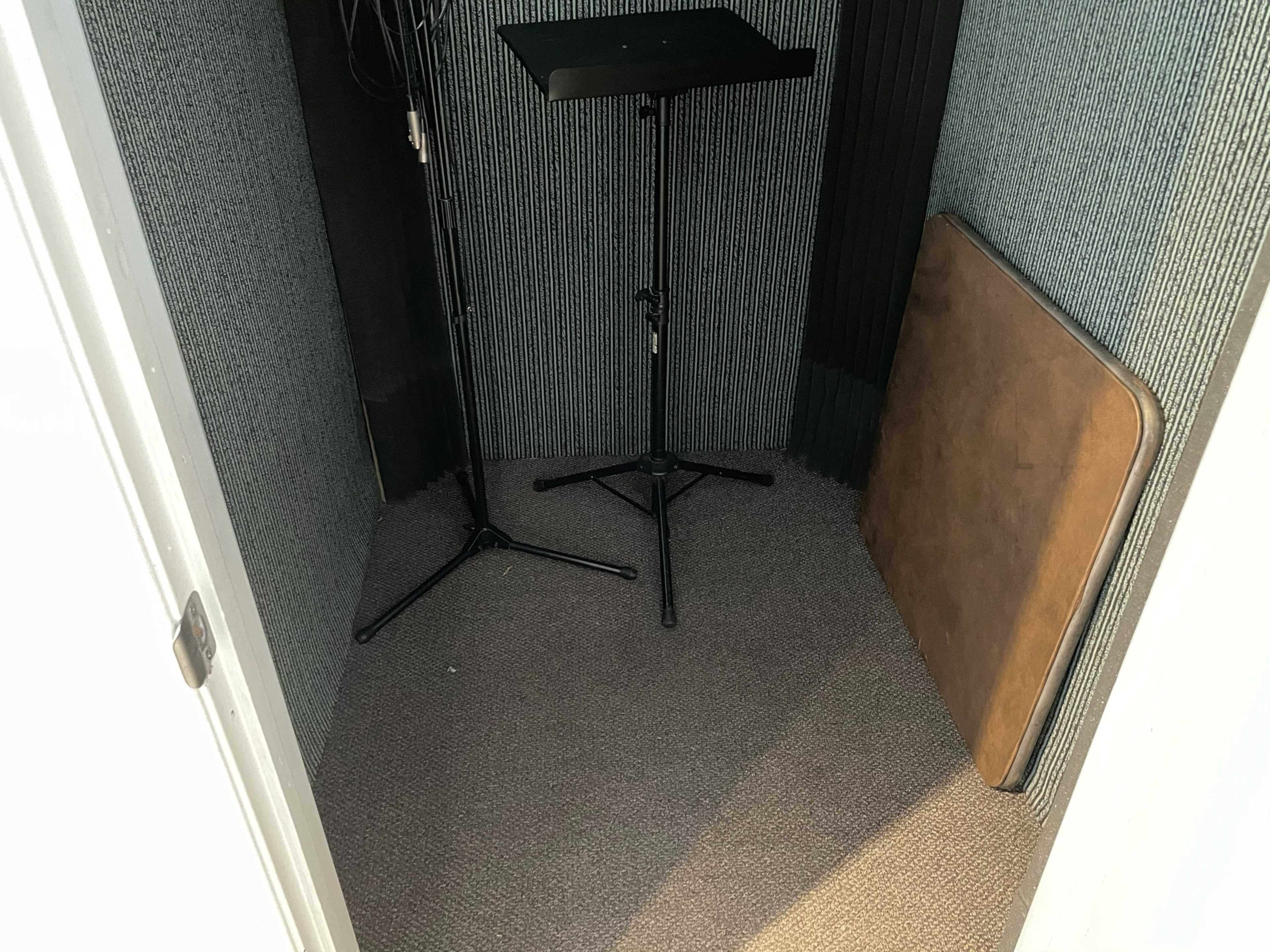 The image shows a small soundproof booth with two microphone stands and a folded table against a carpeted floor.