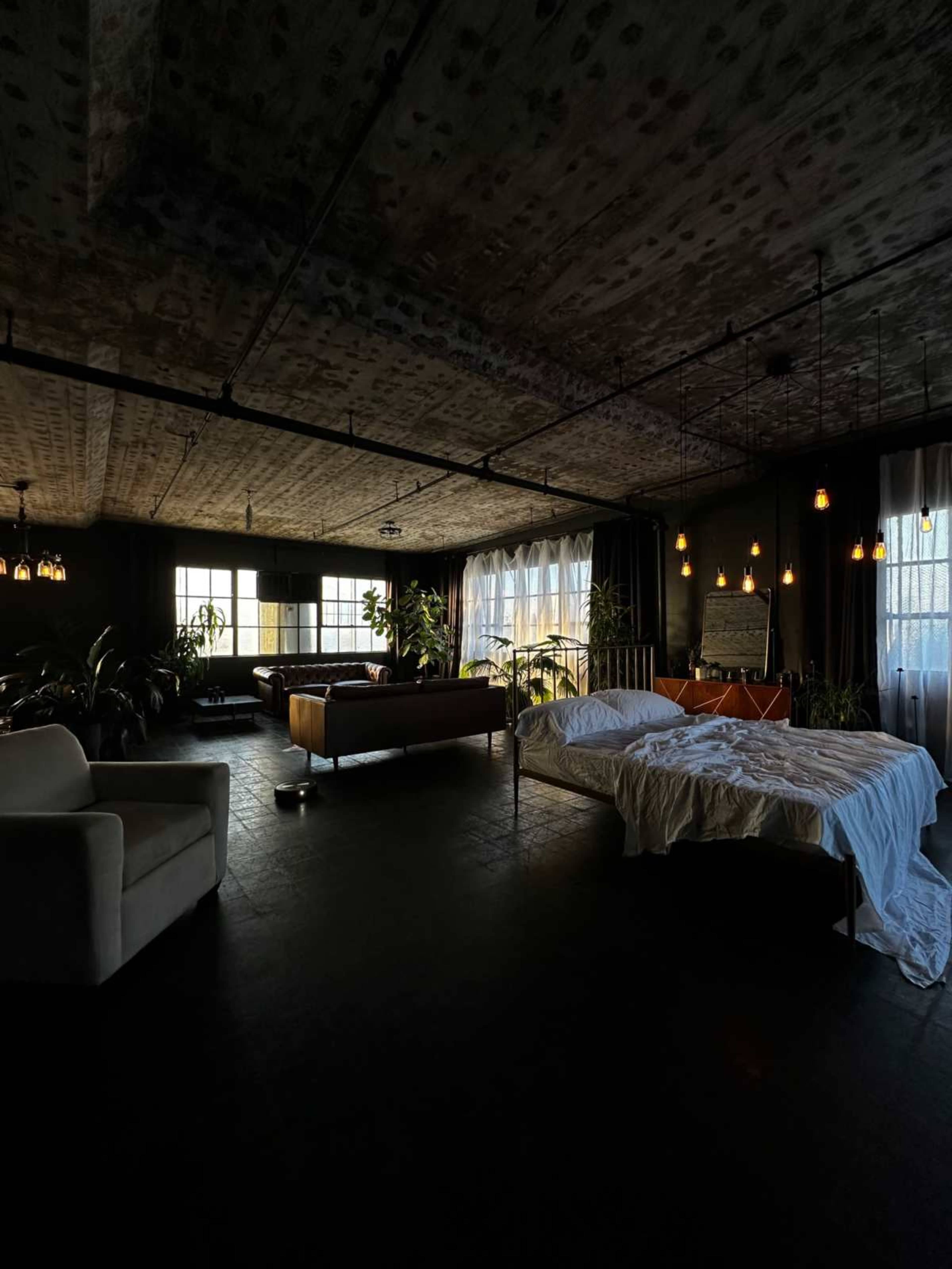 The image shows a spacious, partially furnished room with large windows, plants, and a bed covered with white linens.