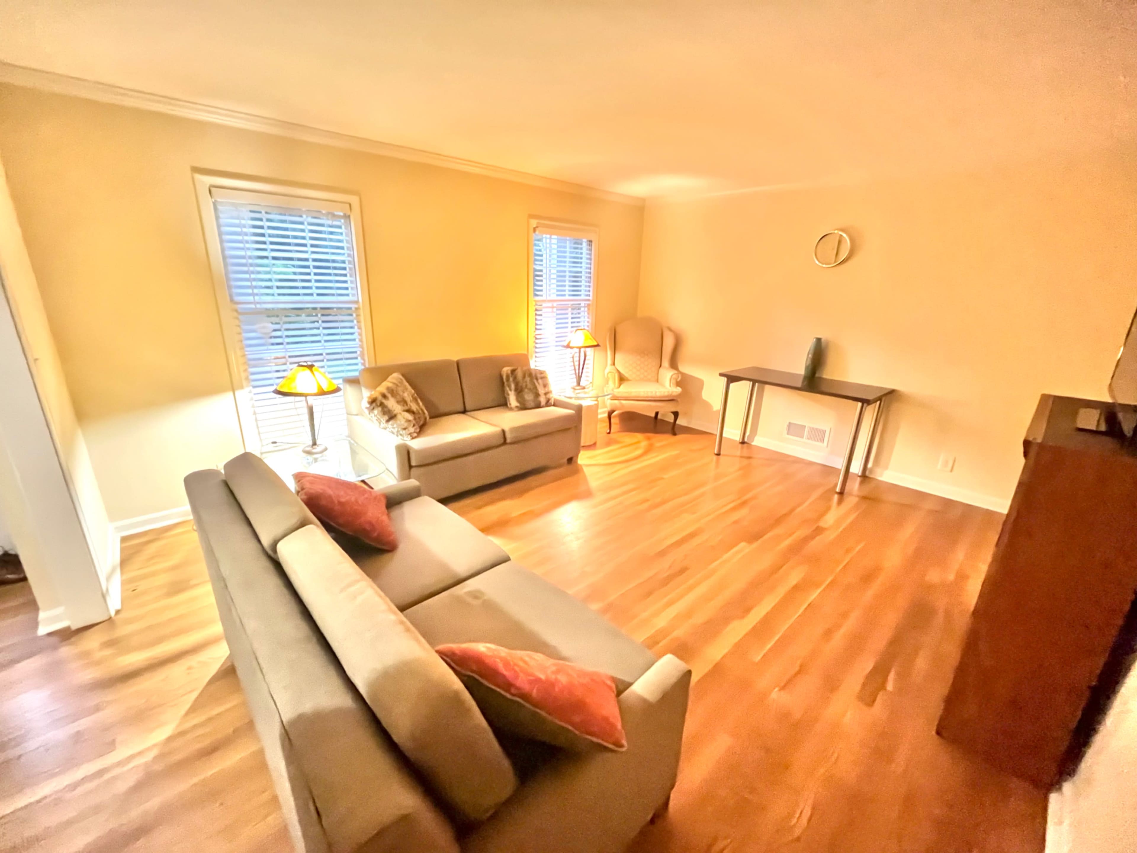 The image shows a cozy living room with hardwood floors, a sofa, an armchair, a small table, and two windows allowing natural light to enter.