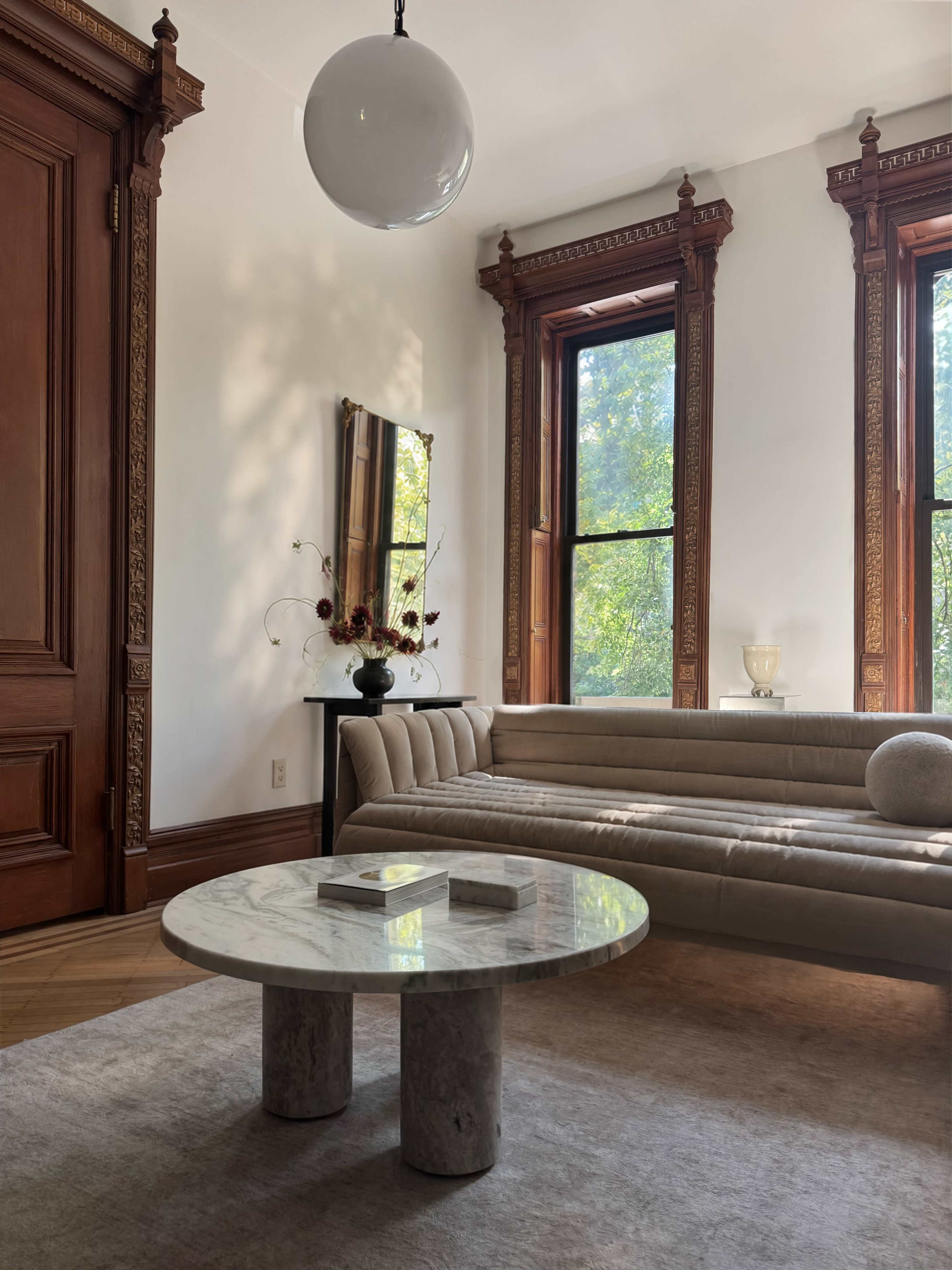 The image shows a bright living room with a marble coffee table, a beige curved sofa, a decorative mirror, and large windows framed by ornate woodwork.