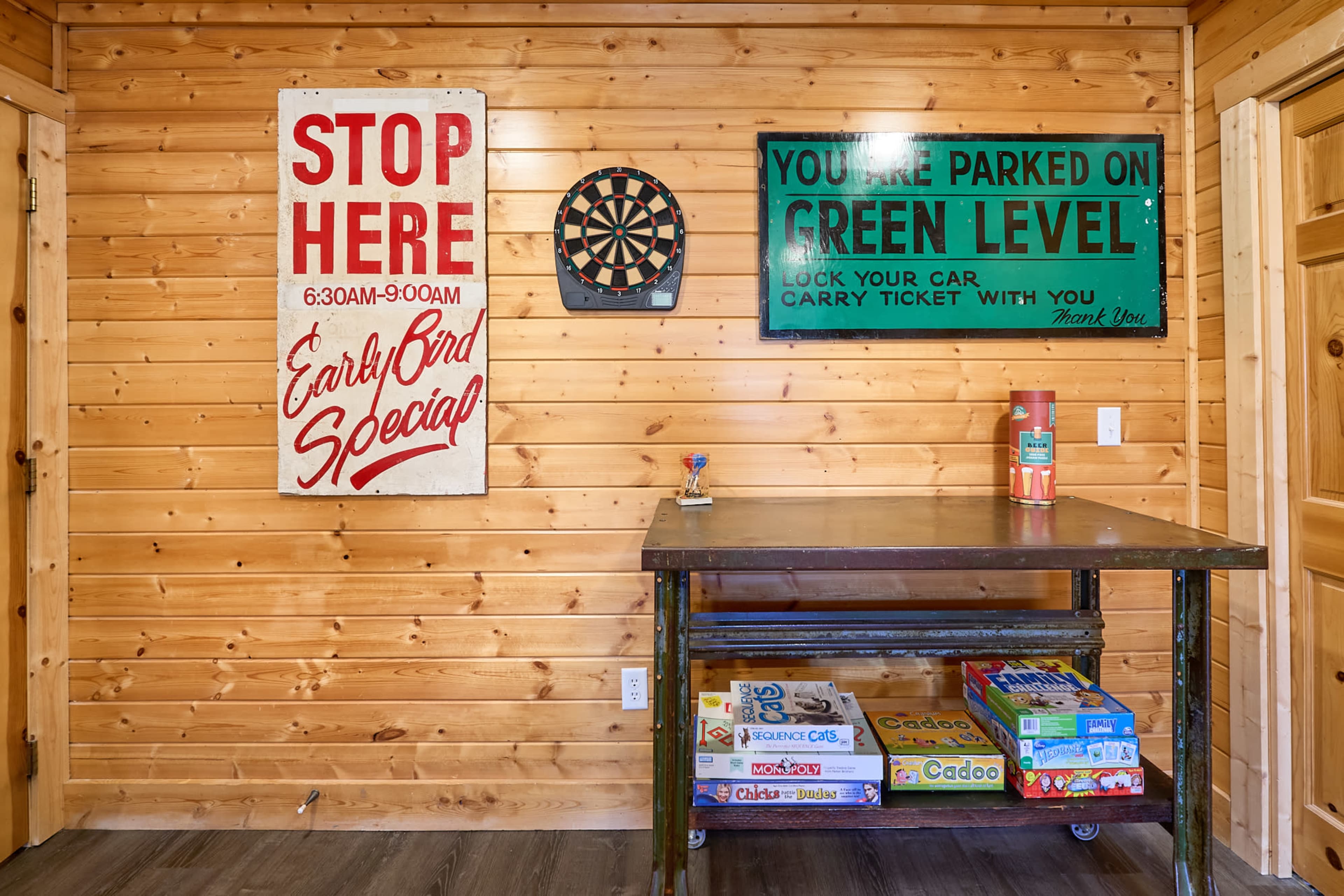 The image shows a wooden-paneled room with two signs on the wall, a dartboard, and a table displaying board games.