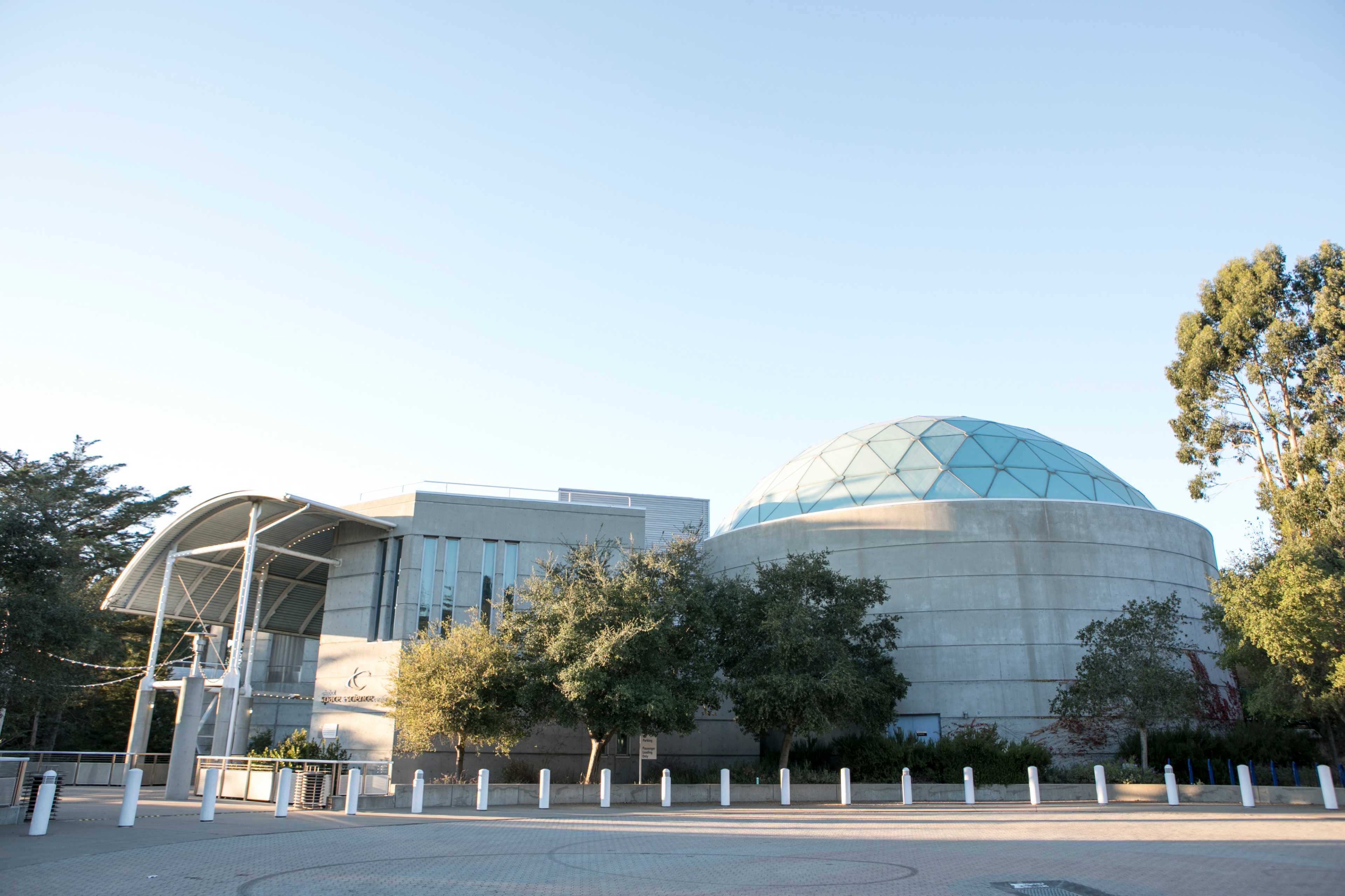 A modern building featuring a geodesic dome and a curved roof, surrounded by trees and a paved area.