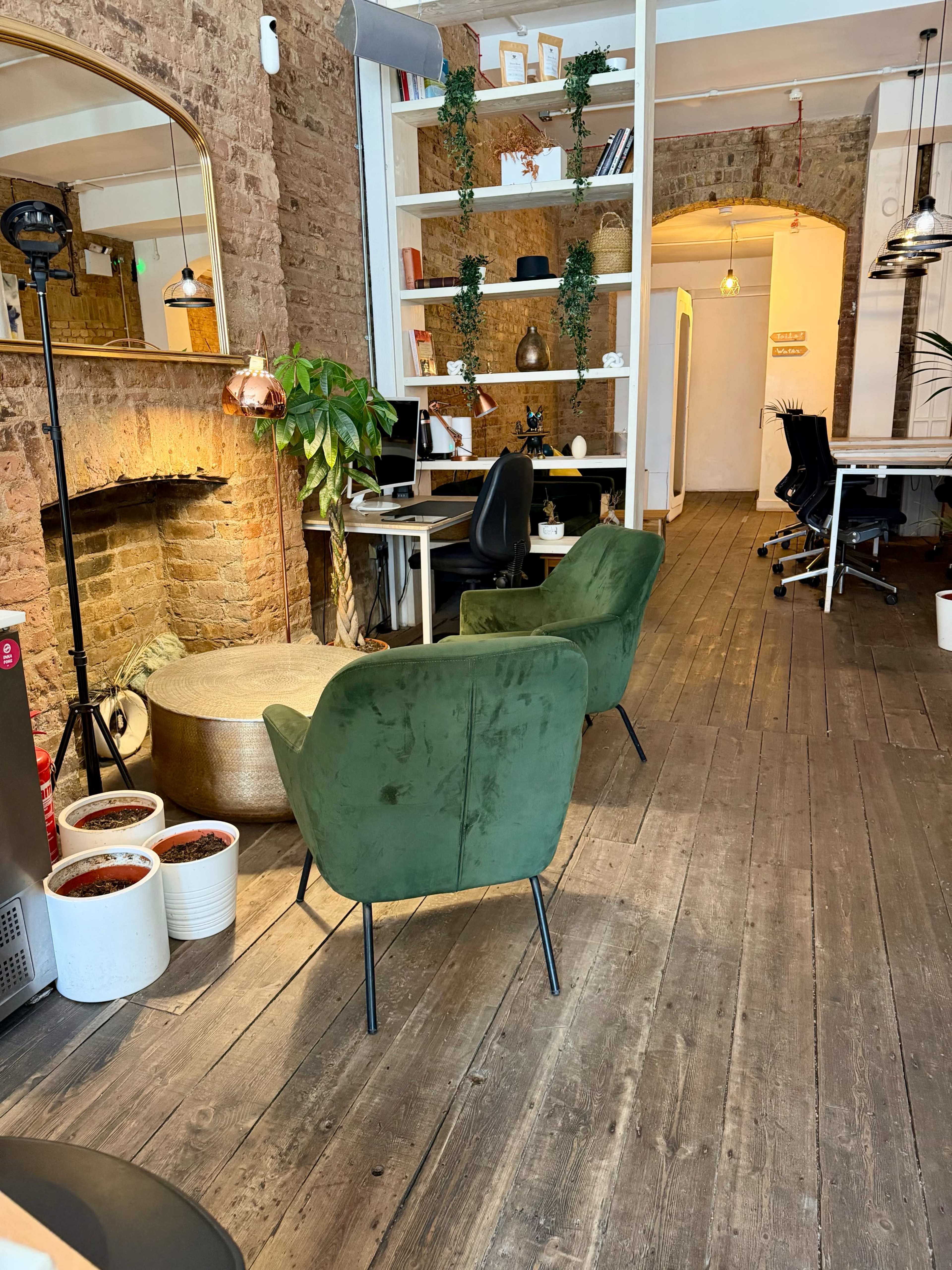 The image shows a modern workspace featuring green chairs, a desk setup, potted plants, and a brick wall with a fireplace.
