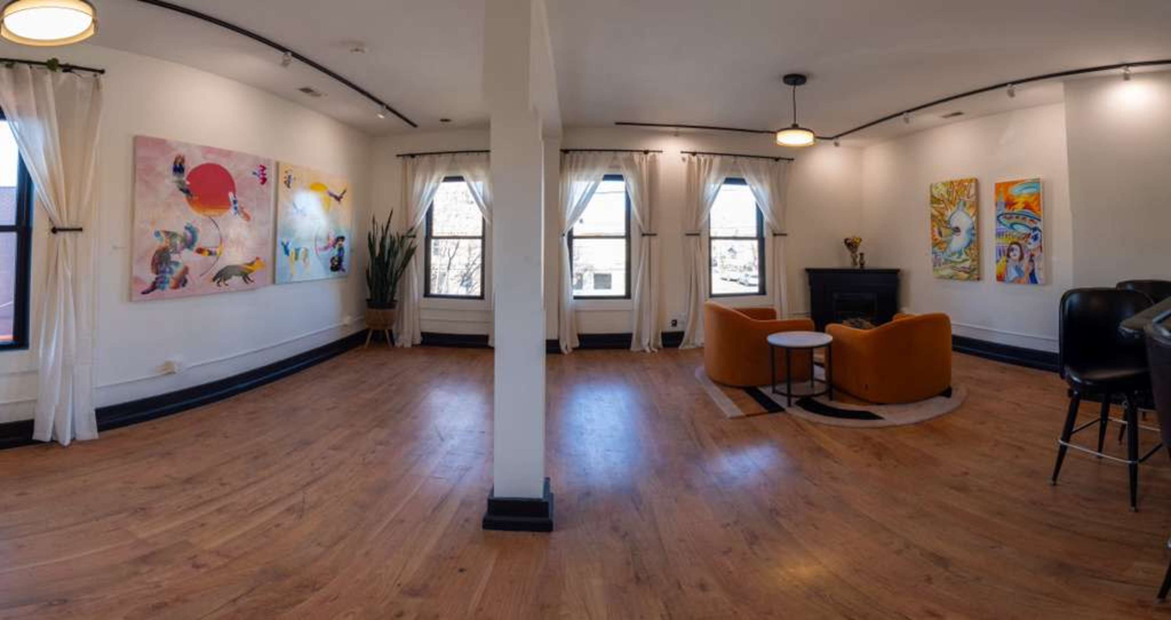 The image shows a well-lit room with large windows, featuring colorful artwork on the walls and a seating area with two orange chairs and a fireplace.