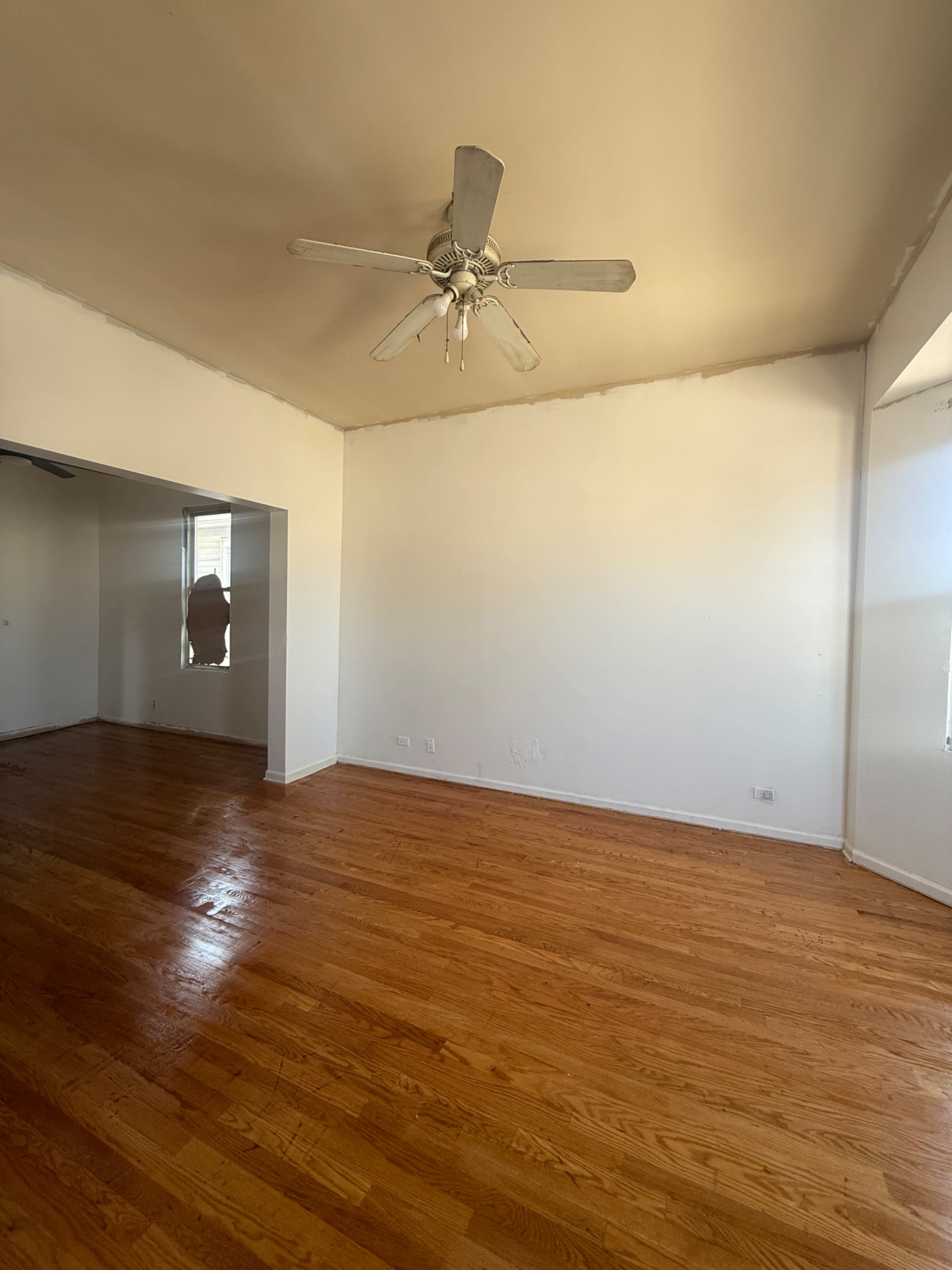 The image shows a room with wooden flooring, a ceiling fan, and plain white walls, without furniture or other decor.