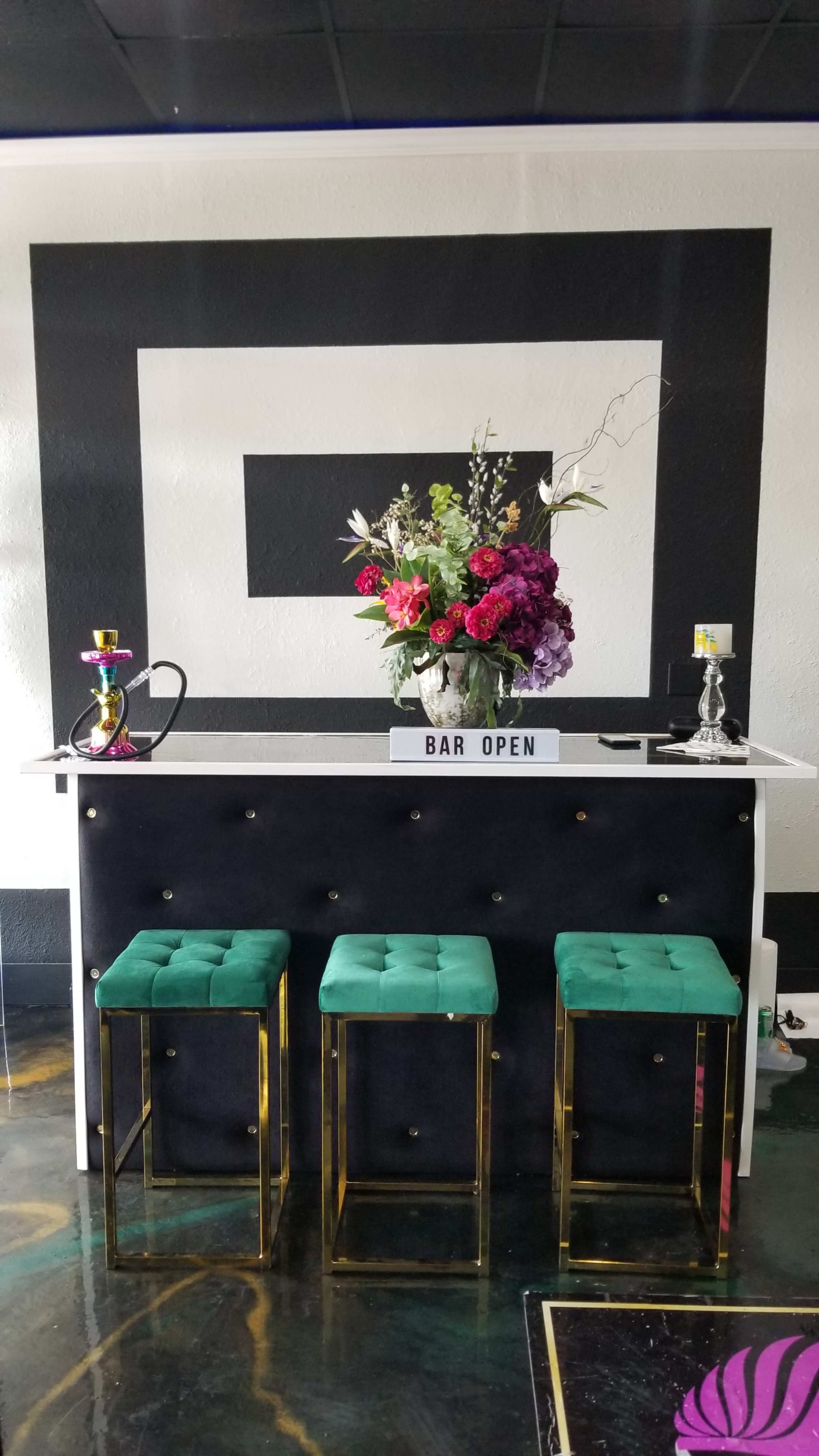 A modern bar area with a black and white geometric design, three green velvet stools, and a floral arrangement on the counter.