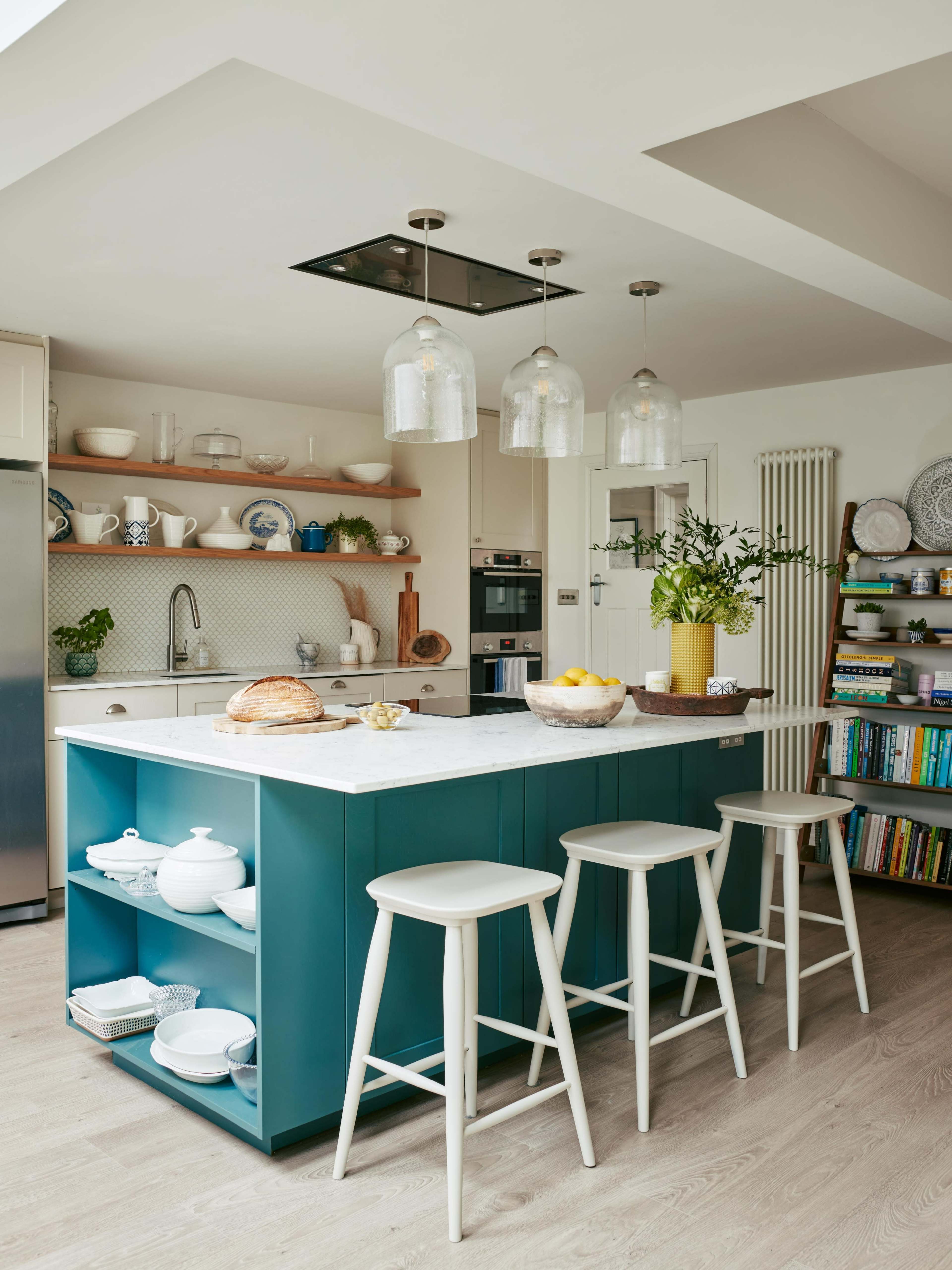 A modern kitchen features a blue island with a marble countertop, three white stools, and open shelving displaying dishes and decor.