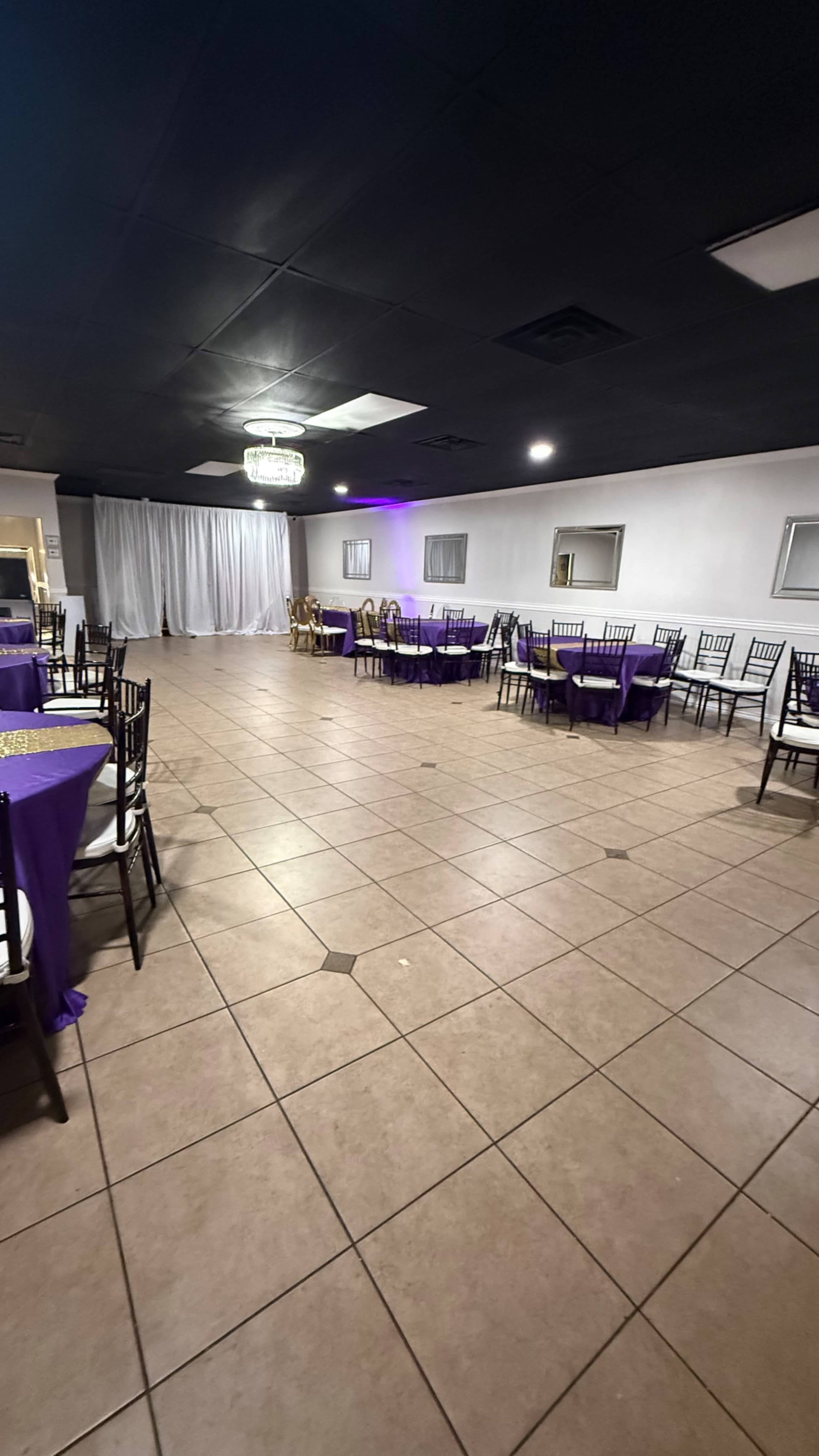 The image shows an empty event space with tables set in a symmetrical layout, decorated in purple, and illuminated by overhead lights.