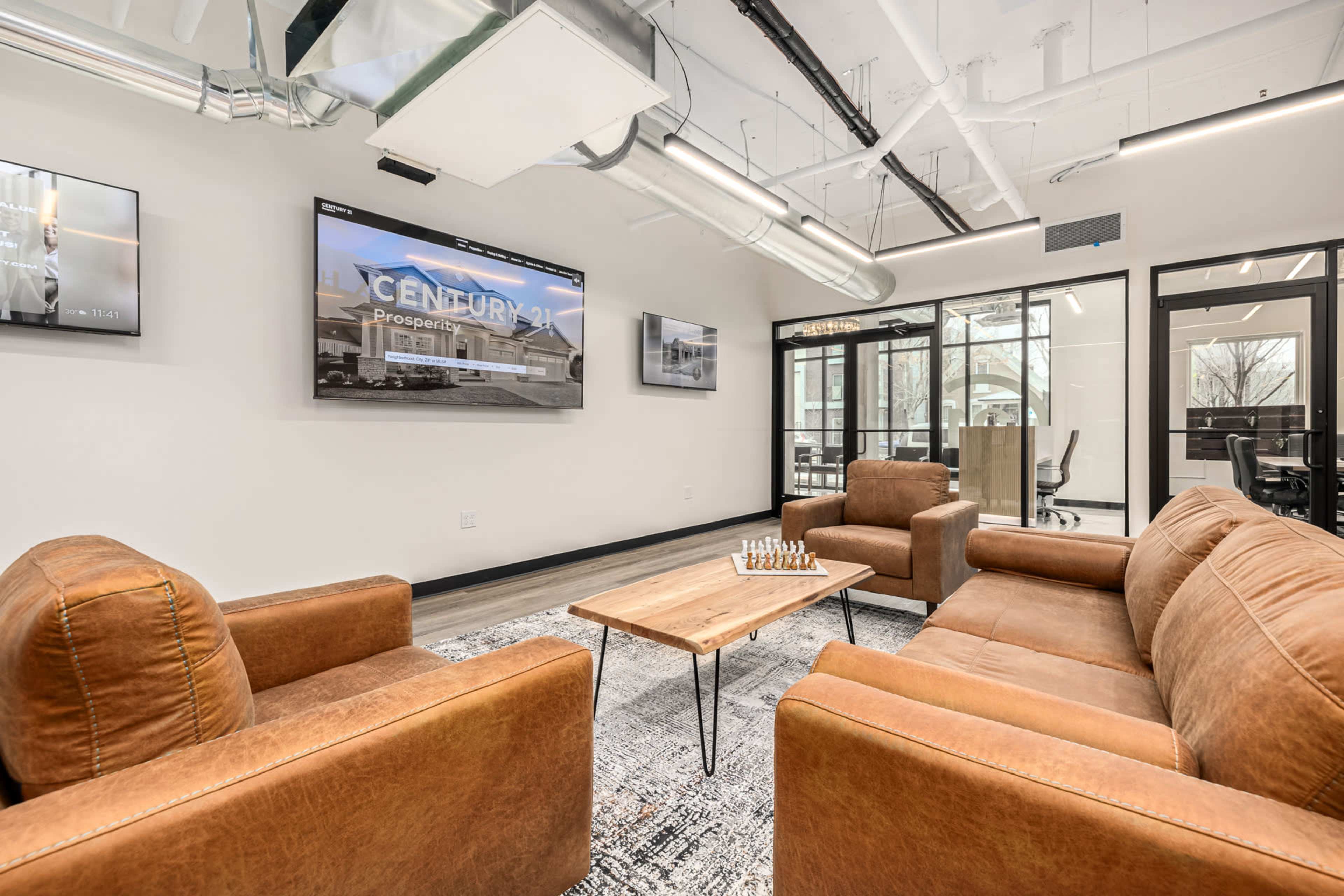 The image shows a modern office lounge area featuring two brown leather chairs, a wooden coffee table with a chess set, and multiple screens displaying images on the walls.