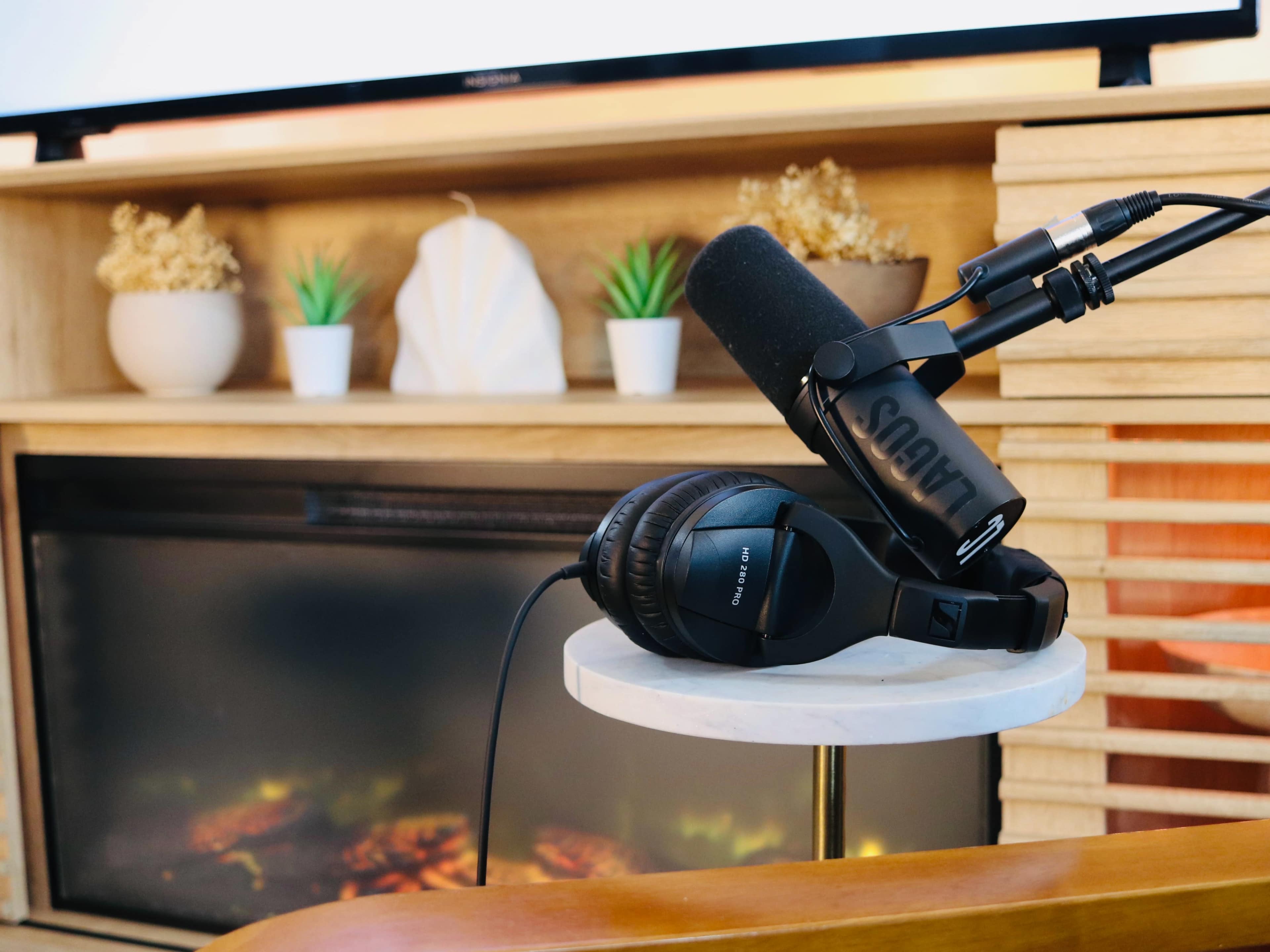 A microphone and headphones rest on a white marble side table in front of a modern fireplace with decorative plants and a television in the background.