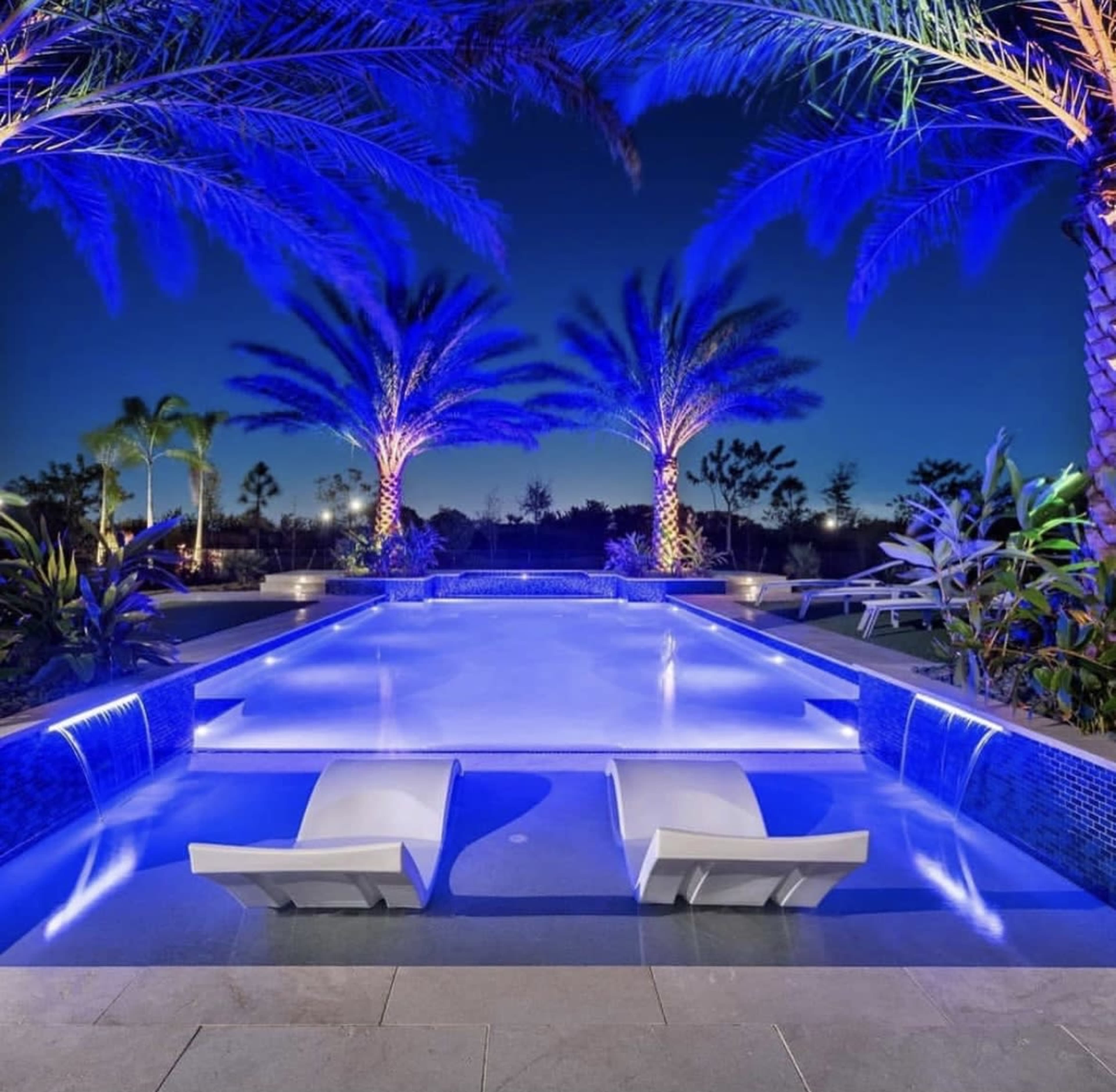 A modern swimming pool with lounge chairs is illuminated by blue lights under palm trees at night.