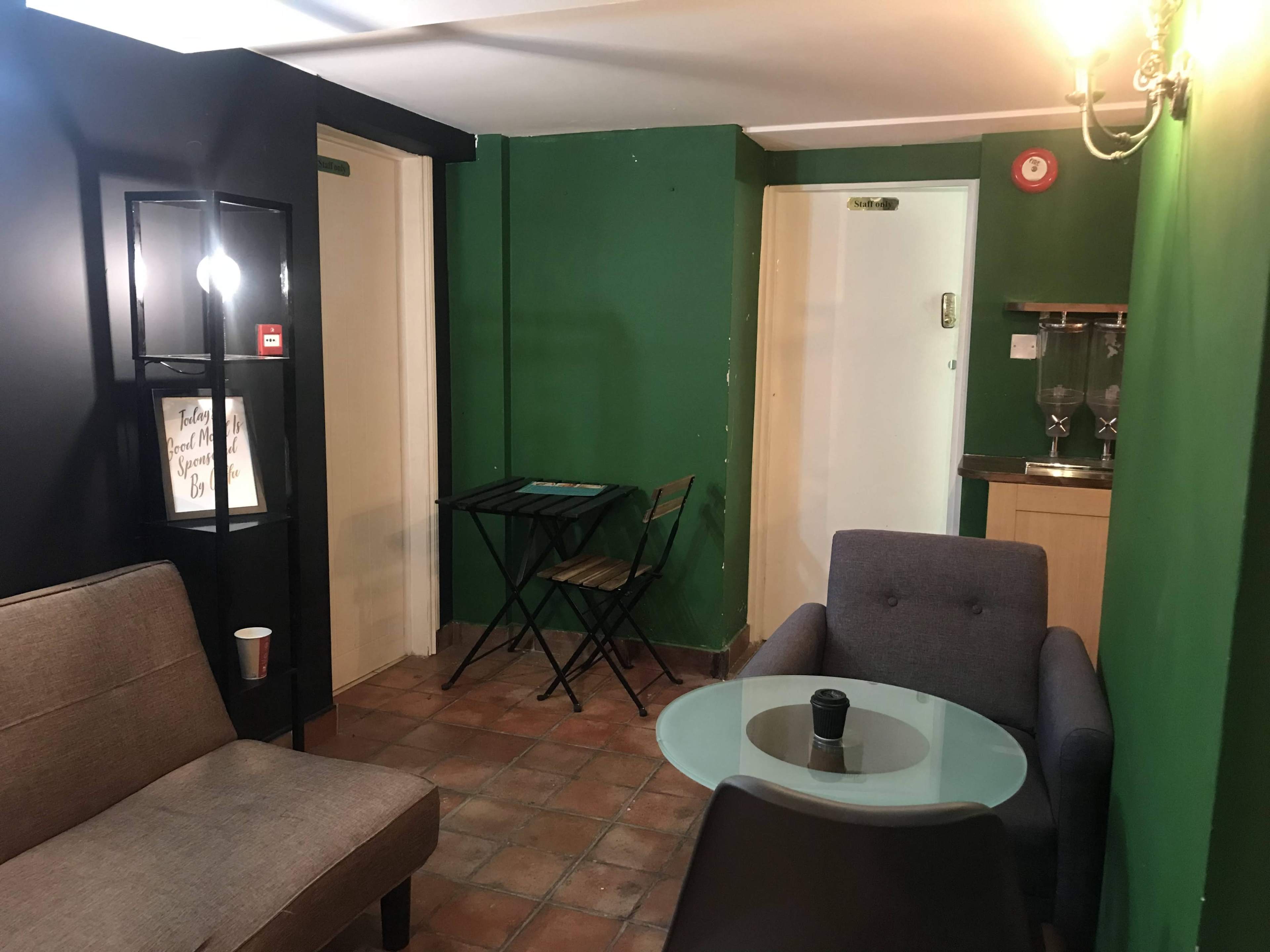 A small lounge area with a gray sofa and armchair, a glass coffee table, a black and gold decorative lamp, and a compact dining table against a green wall.