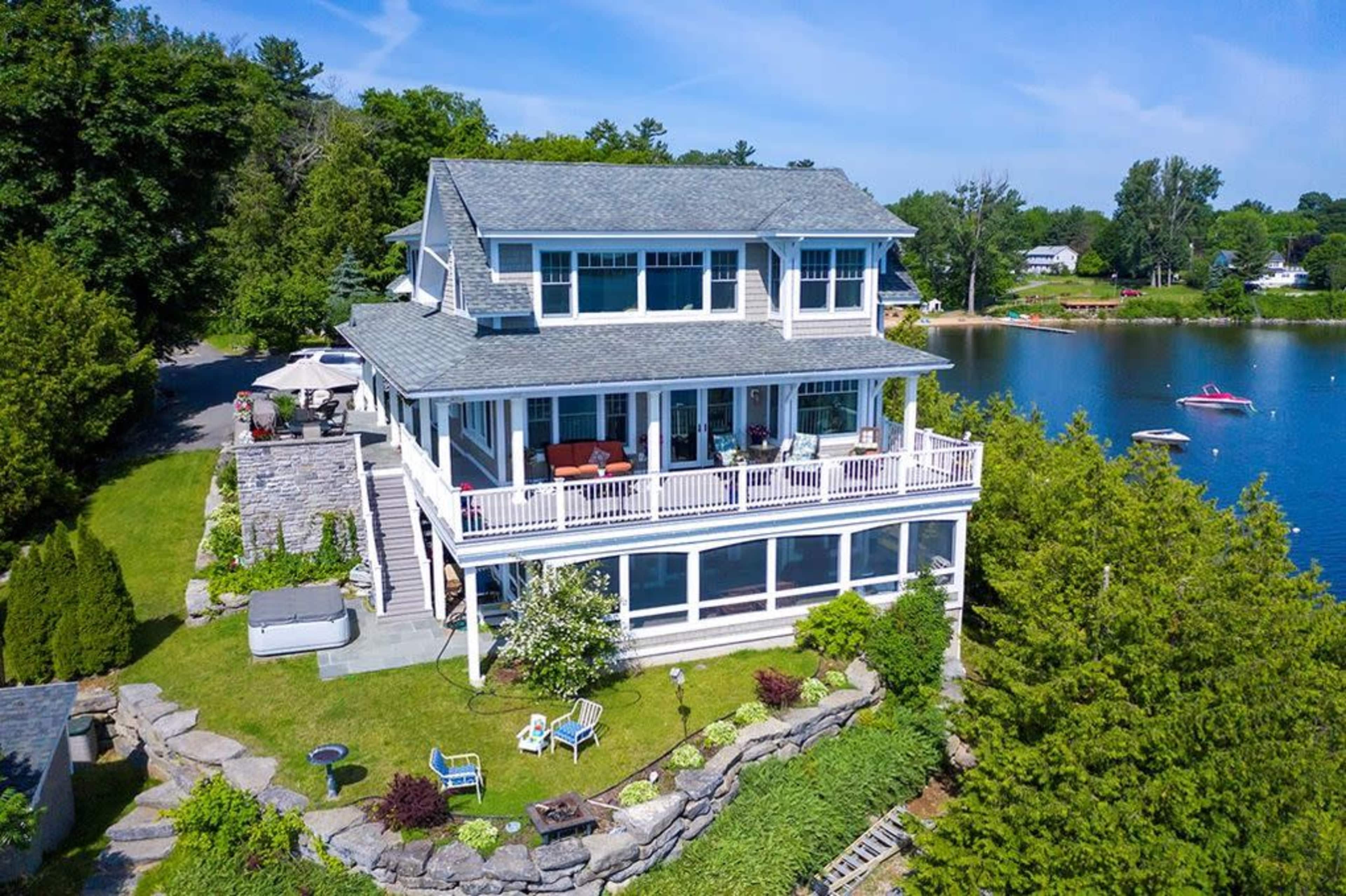 A large, multi-story house beside a lake, featuring a wrap-around porch and landscaped yard with seating areas.