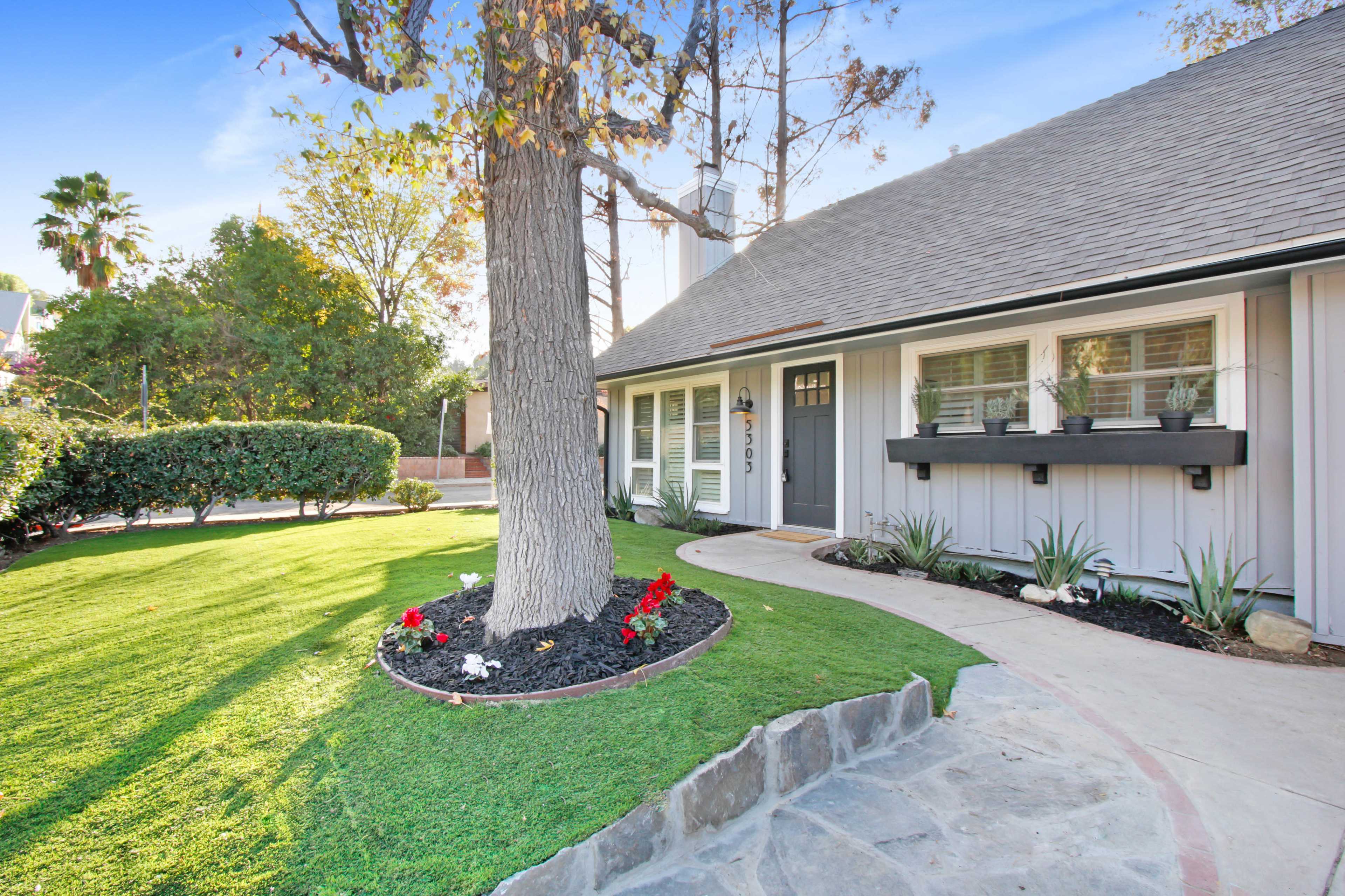 A house with a gray exterior, a front walkway leading to the door, and a landscaped yard featuring a tree and flower beds.