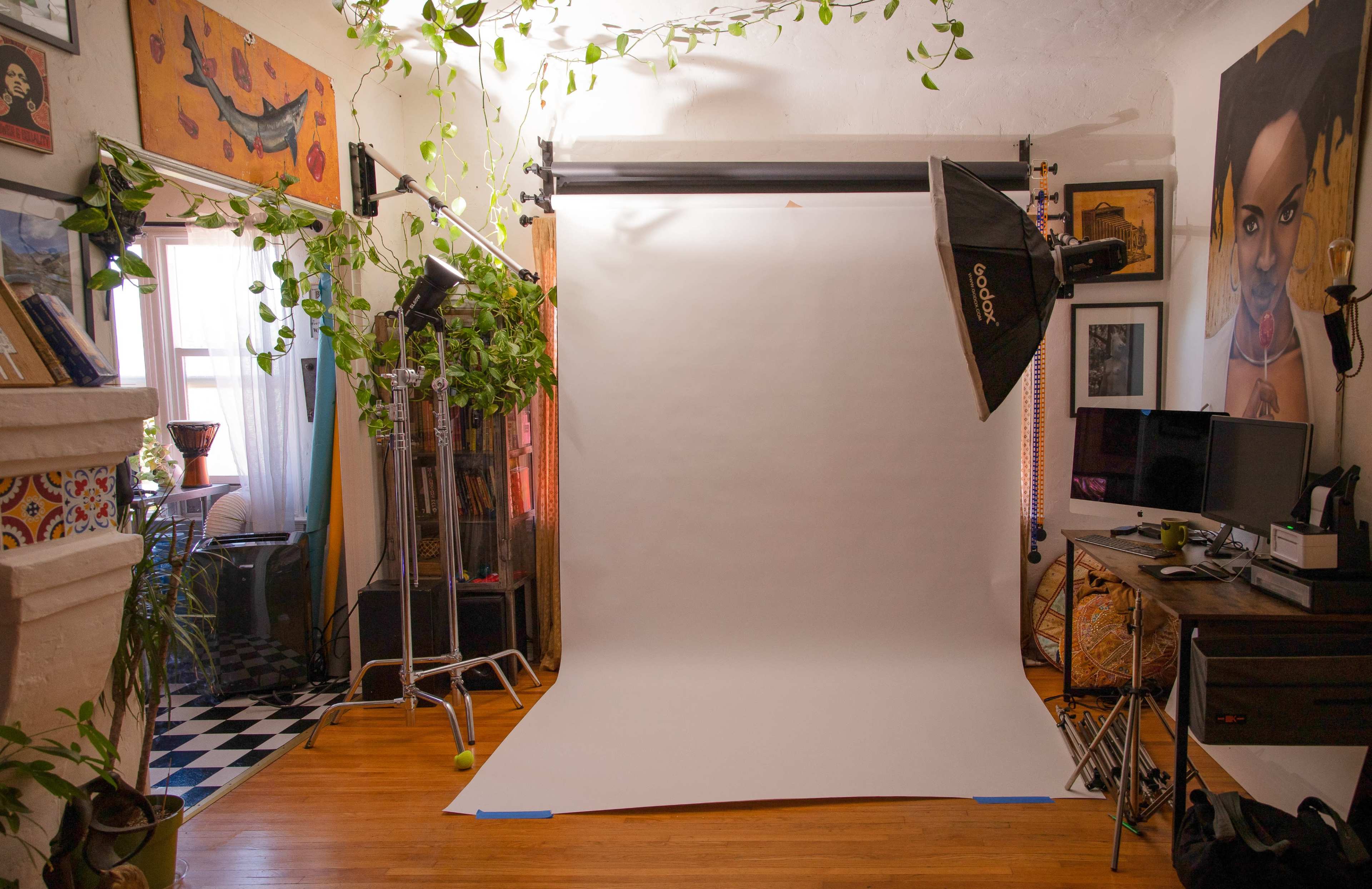 A photography setup in a room with a white backdrop, studio lights, and potted plants, alongside a computer and artwork on the walls.