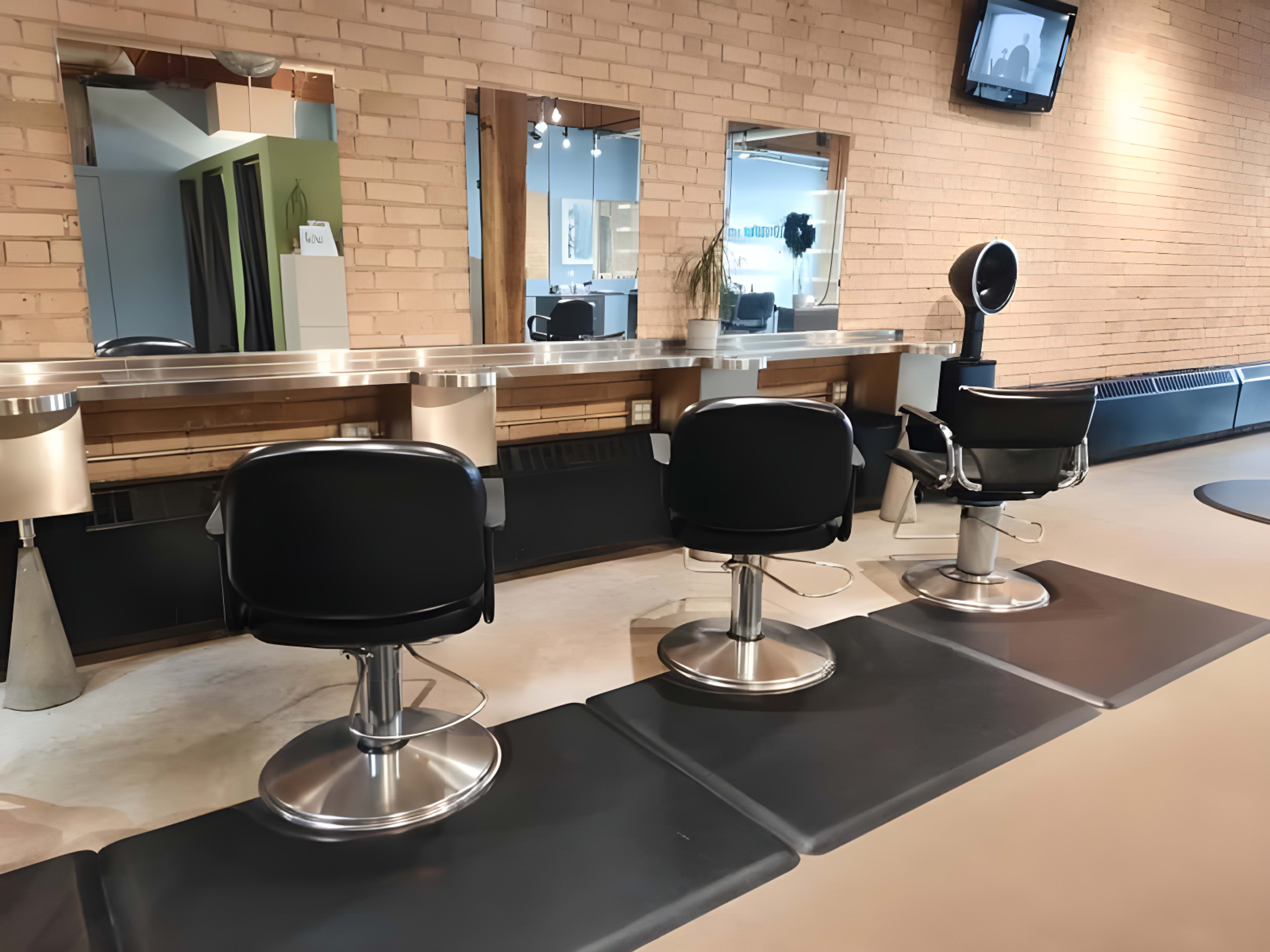 A modern salon interior with three black styling chairs arranged in front of a set of mirrors against a brick wall.