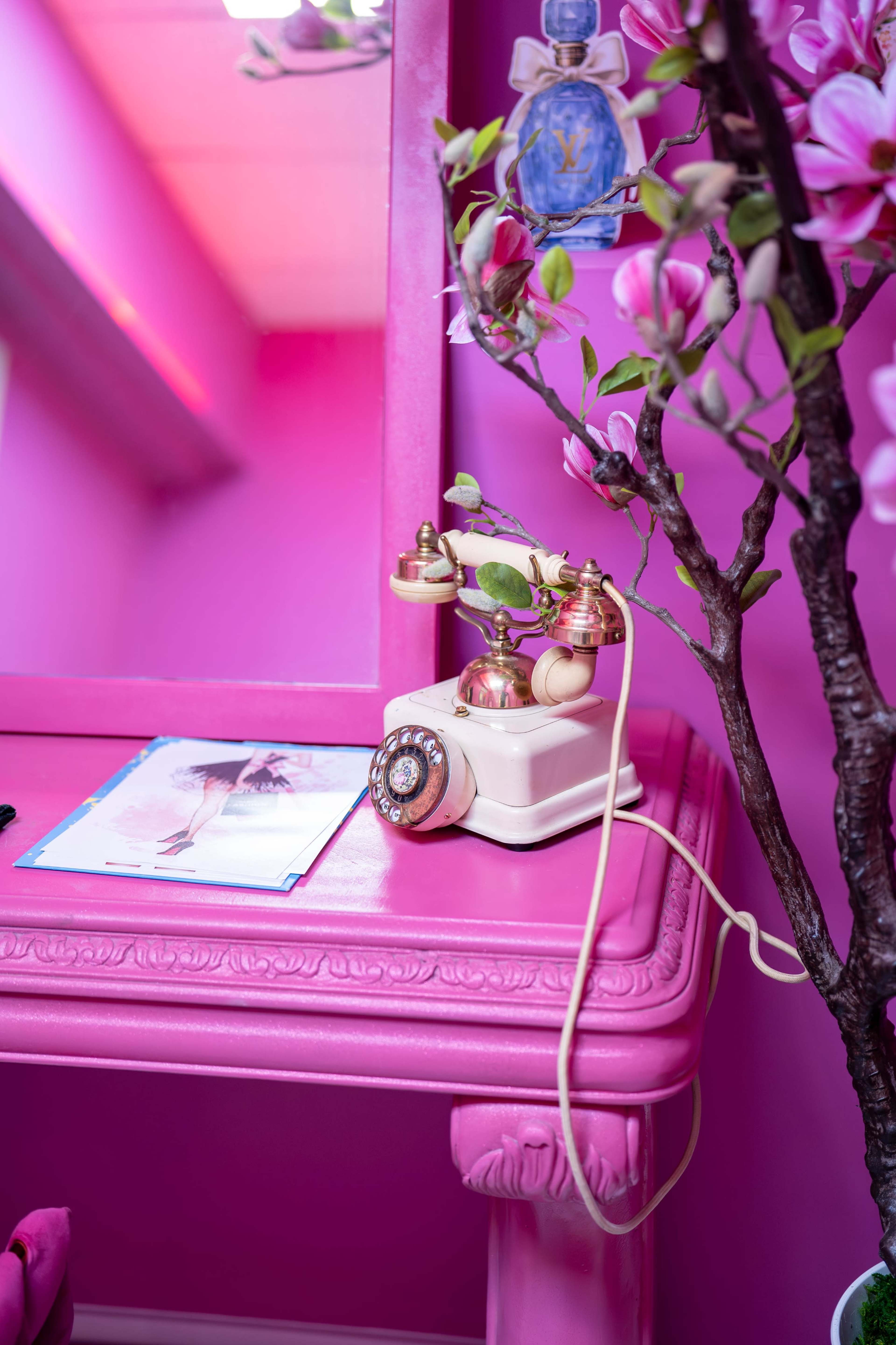 A vintage telephone sits on a pink table next to a magazine and a decorative floral arrangement.