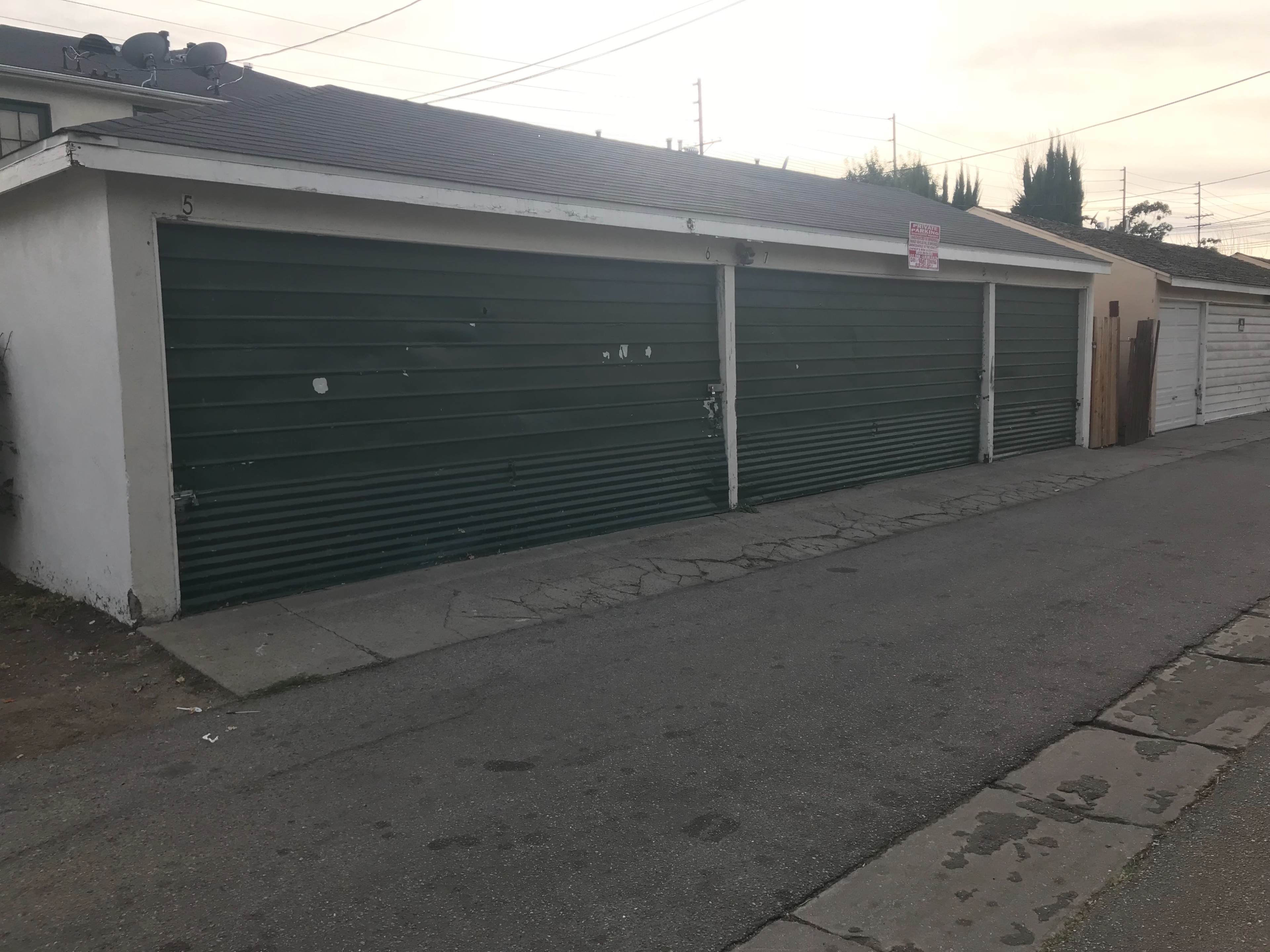 The image shows a row of three green garage doors along a concrete alleyway with a sign on the first door and a paved surface on the right.