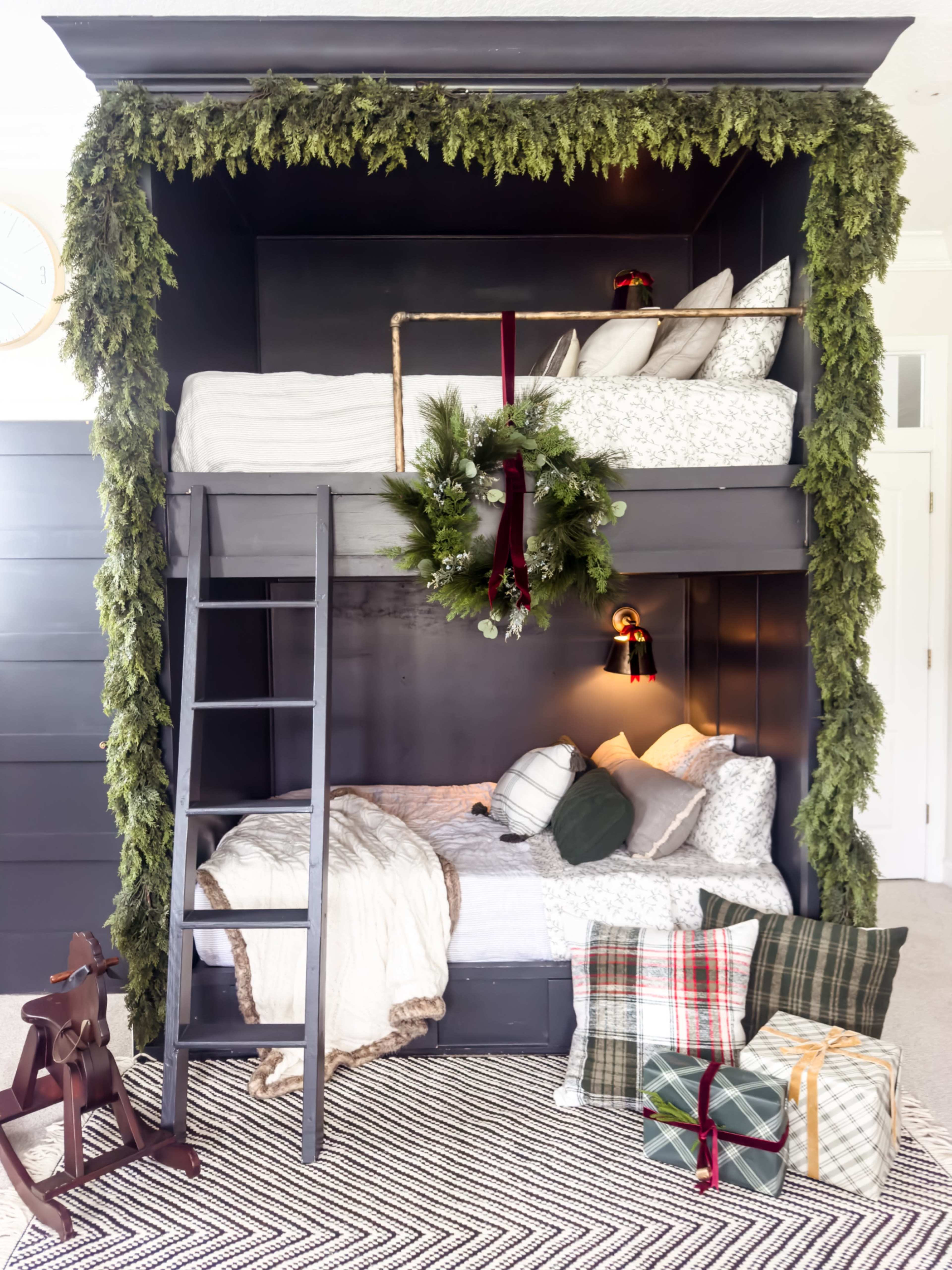 The image features a double bunk bed with a dark wooden frame, adorned with greenery around the top, and flanked by a wreath, while a cozy arrangement of blankets and wrapped gifts is placed on the floor below.