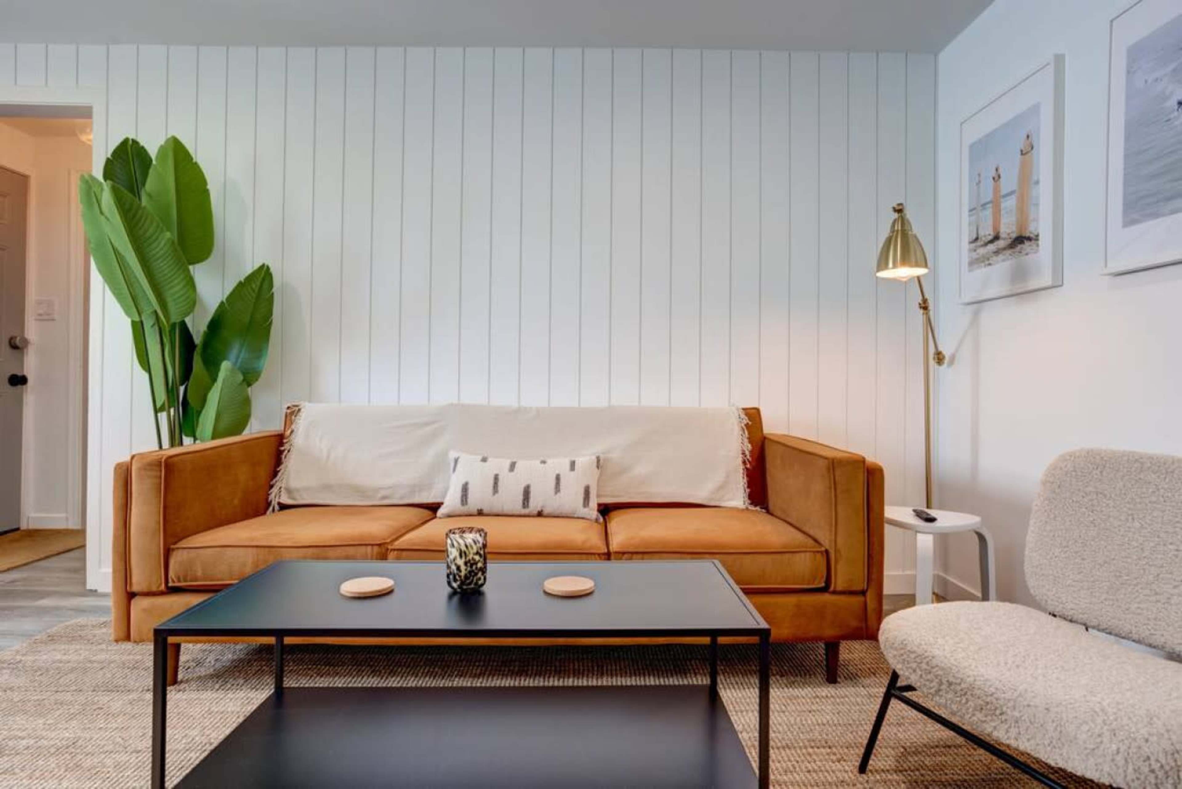 The image shows a modern living room with a tan sofa, a black coffee table, and a potted plant against a white shiplap wall.