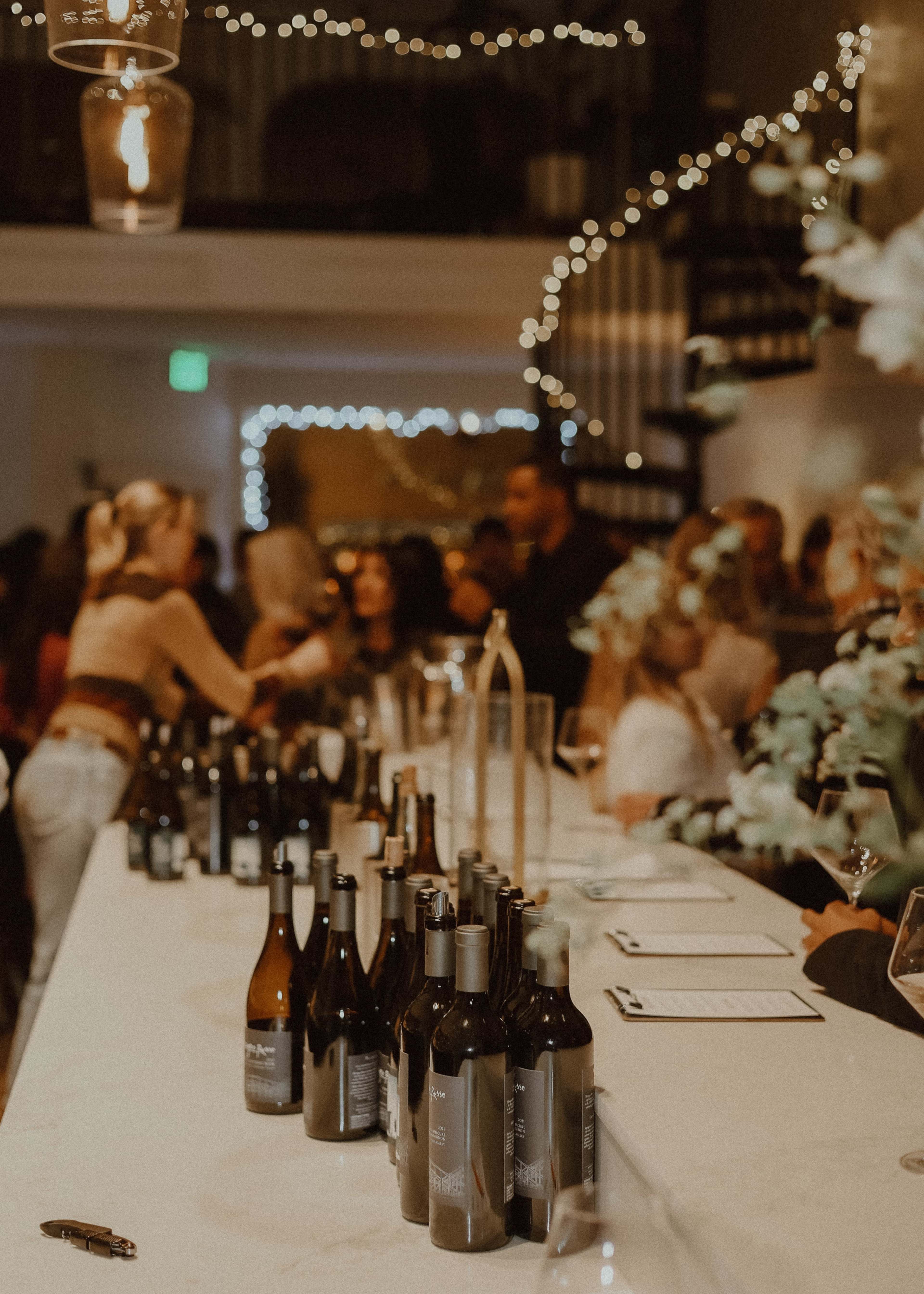 A bar area filled with various wine bottles is bustling with people socializing in a warmly lit setting adorned with string lights.