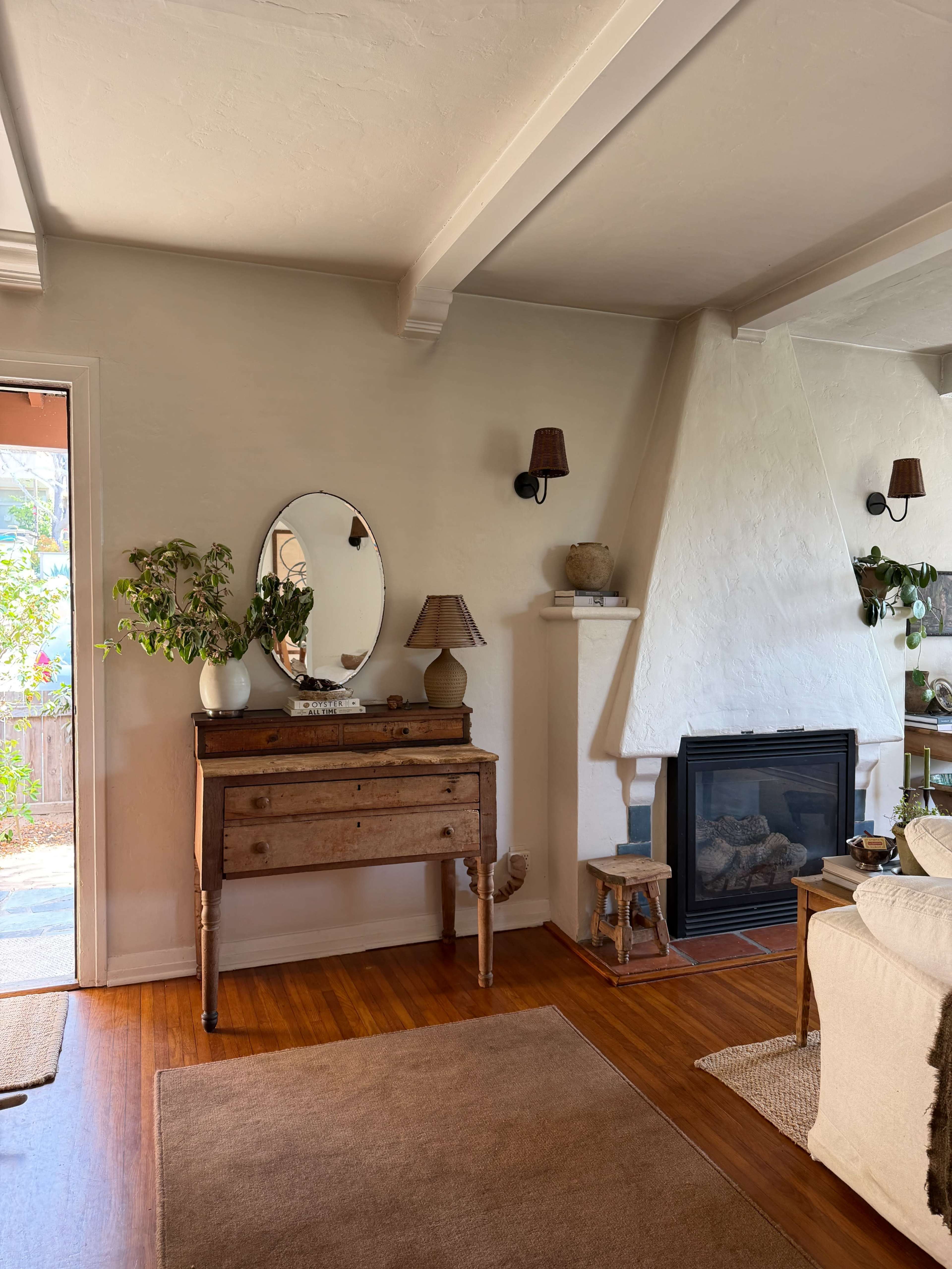 The image shows a cozy living room featuring a wooden console table with a round mirror above it, a fireplace with a stone surround, and warm wooden flooring.