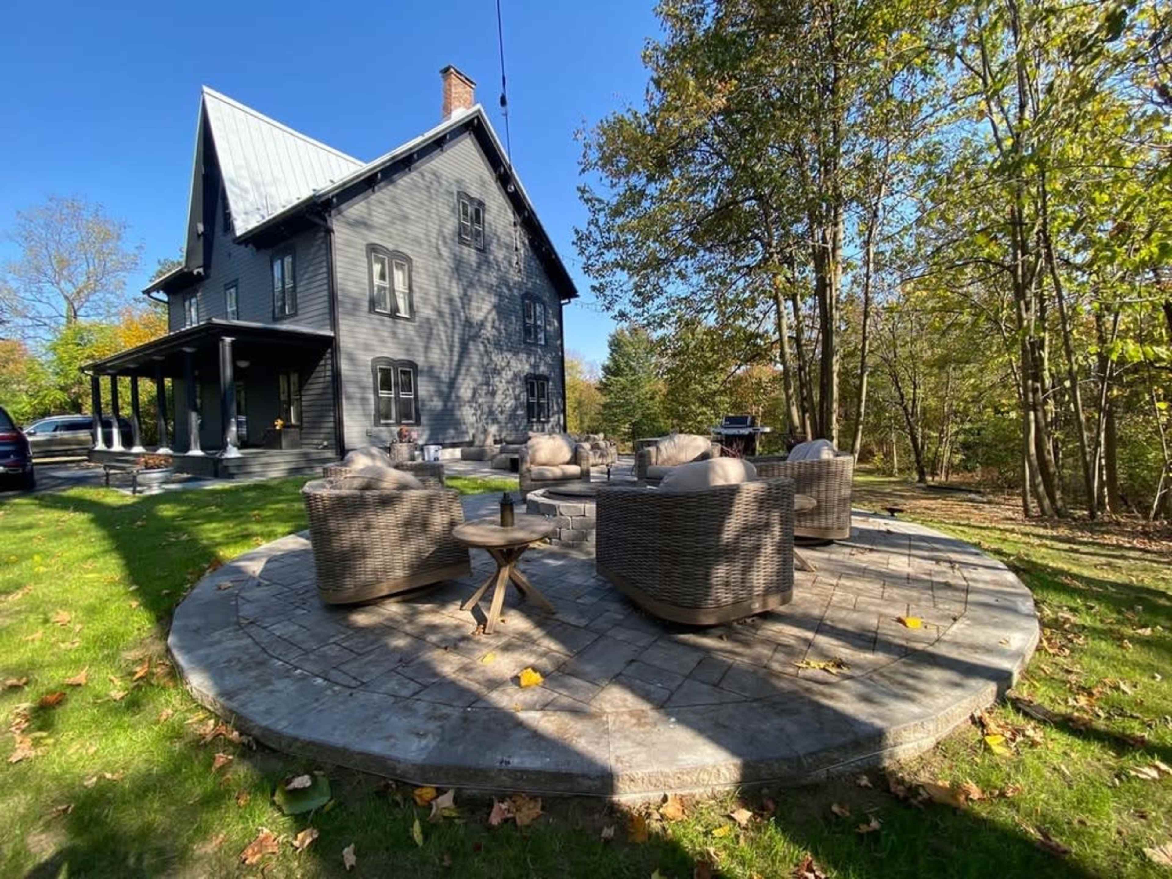 A gray house with a metal roof stands beside a circular stone patio surrounded by outdoor chairs and trees.