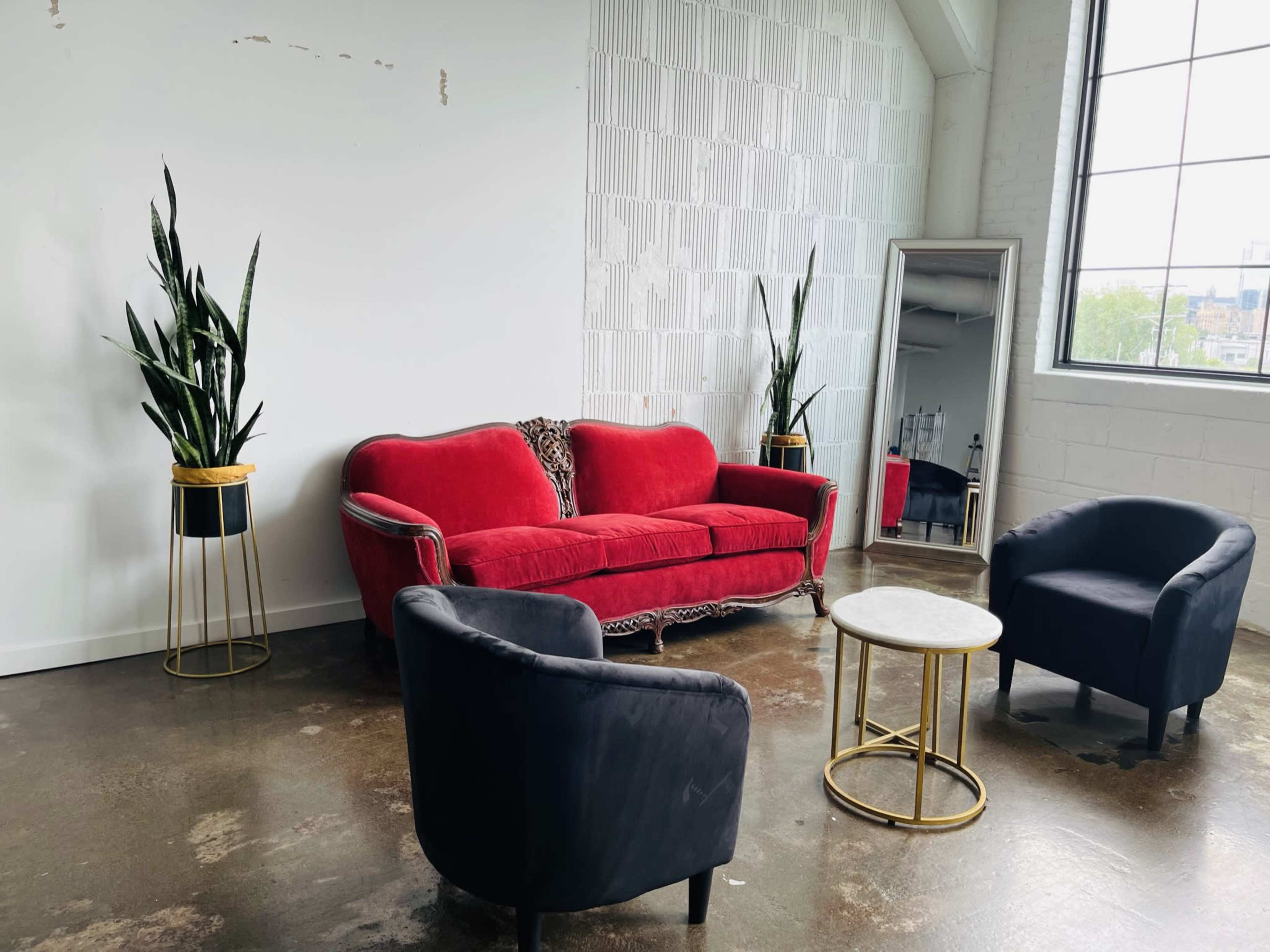 A bright, minimalist room featuring a red vintage sofa, two black armchairs, a round white coffee table, a large mirror, and two potted plants.