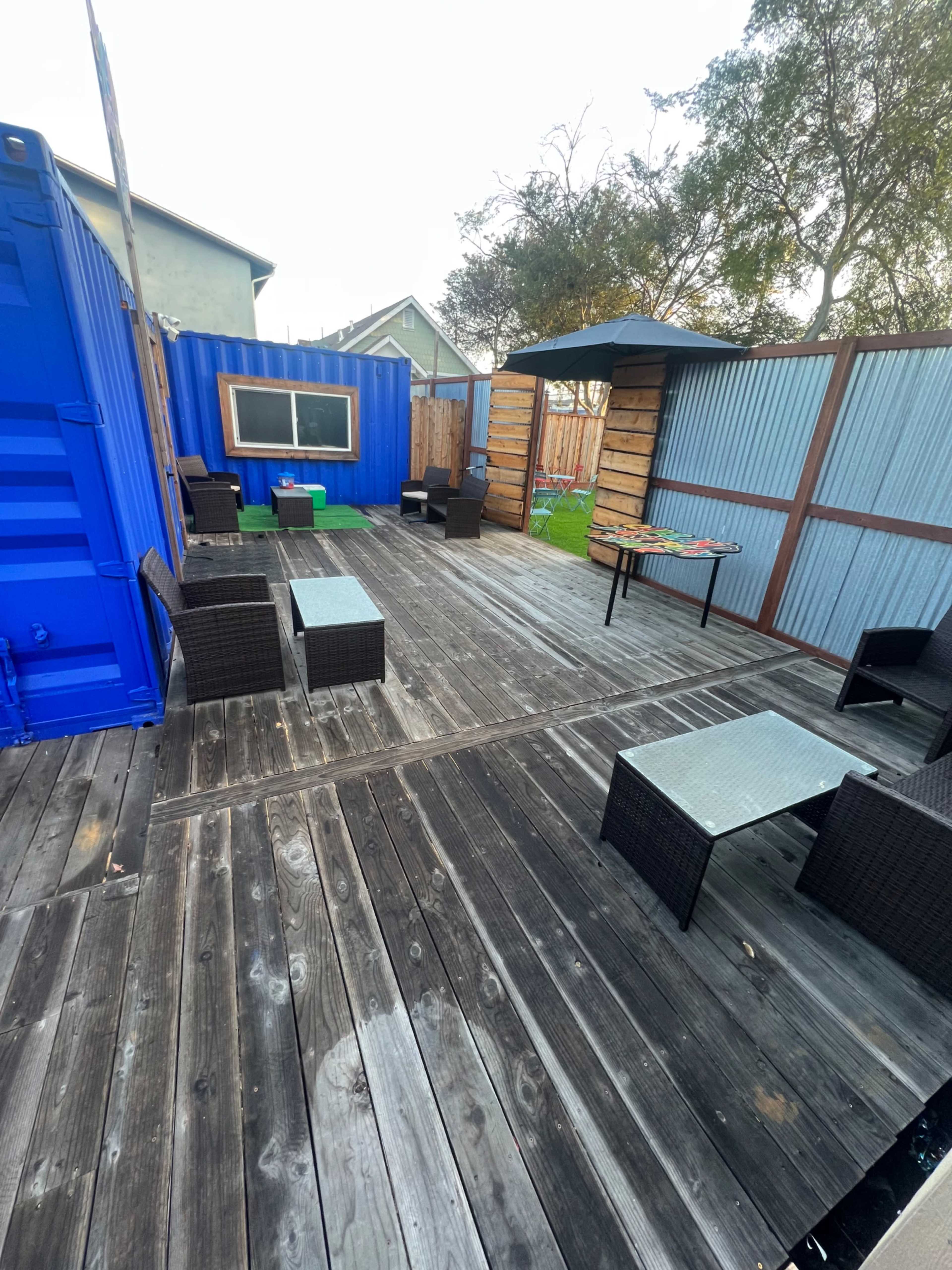 The image shows an outdoor seating area with wooden flooring, featuring several wicker chairs and tables, a large blue shipping container, and a small grassy section.