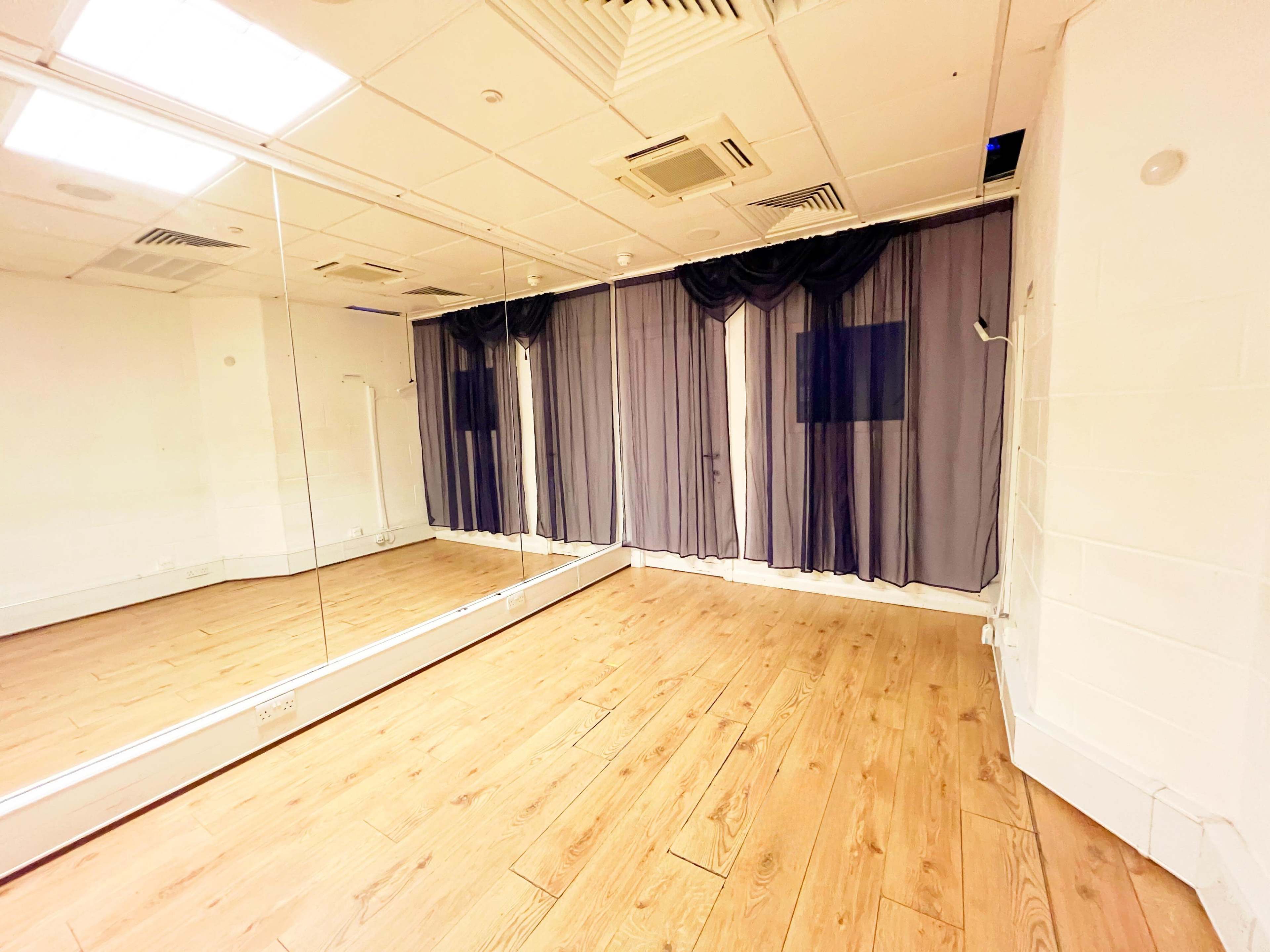 The image shows an empty room with mirrors on one wall, wooden flooring, and sheer curtains covering the windows.